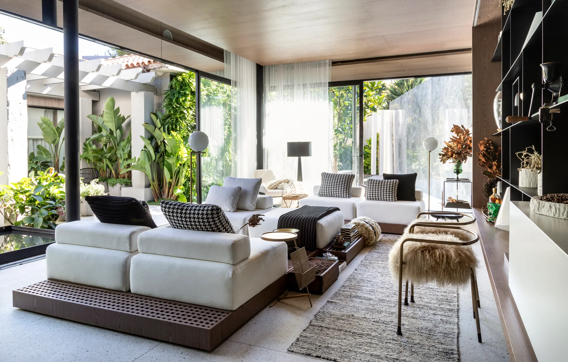 A modern living room features a white modular sofa on a wood base, terrazzo flooring, and glass walls looking out to a lush garden.