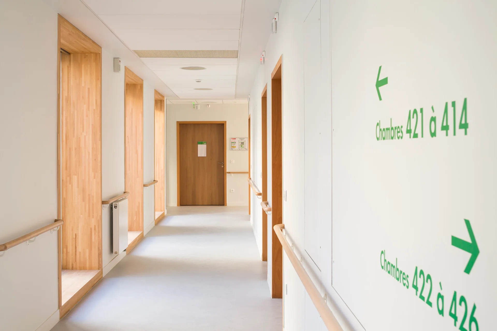 A bright white corridor features recessed wood-lined alcoves, matching light wood handrails, and green directional signage on the wall.