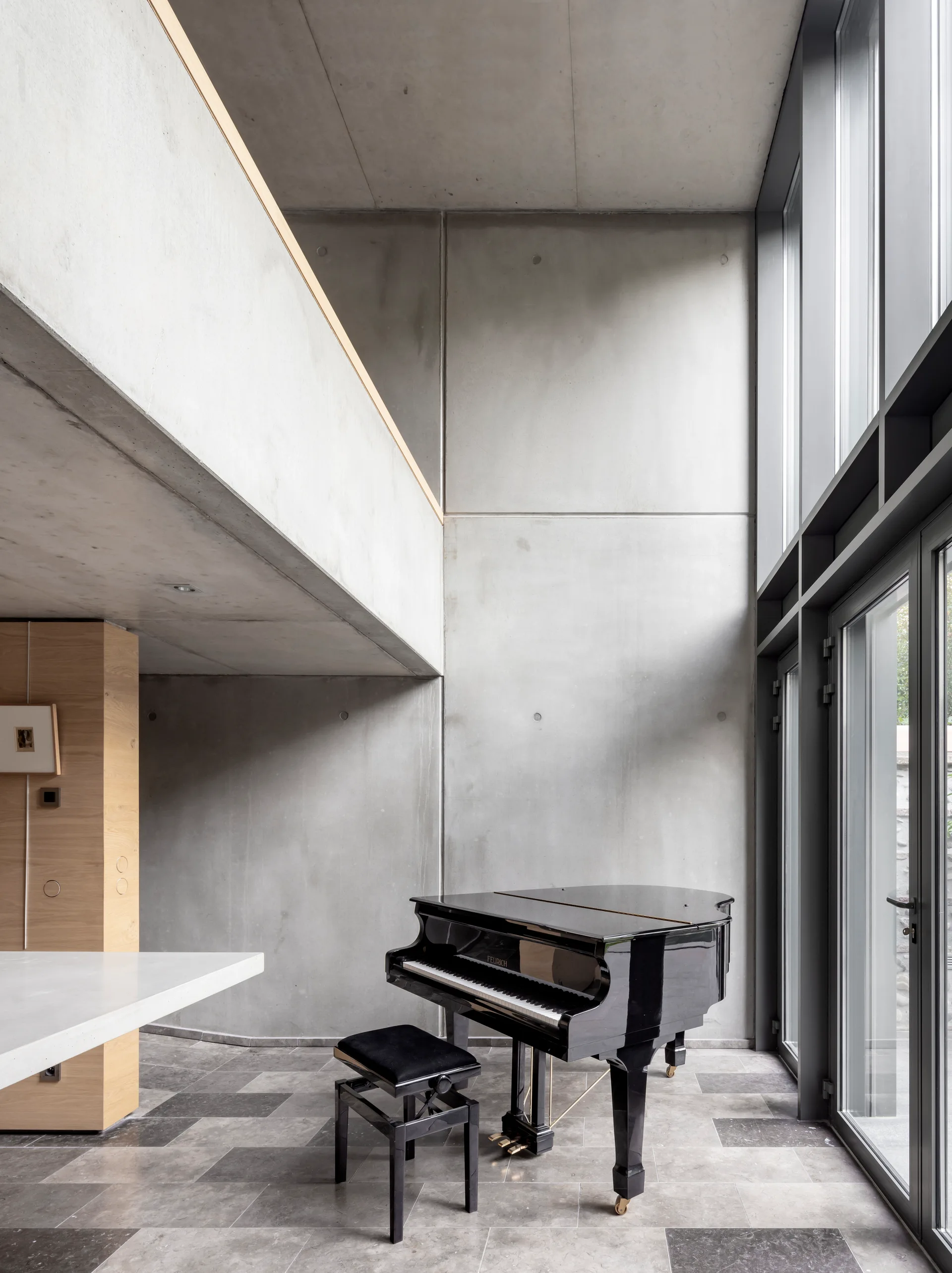 A black grand piano sits on grey stone tiles in a modern room with double-height exposed concrete walls and high windows.