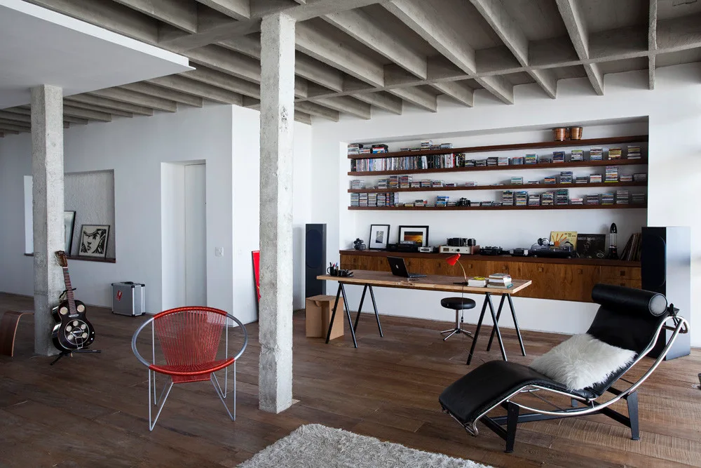 A spacious open living room featuring an exposed concrete waffle ceiling, concrete columns, a desk, and a black leather chaise lounge.