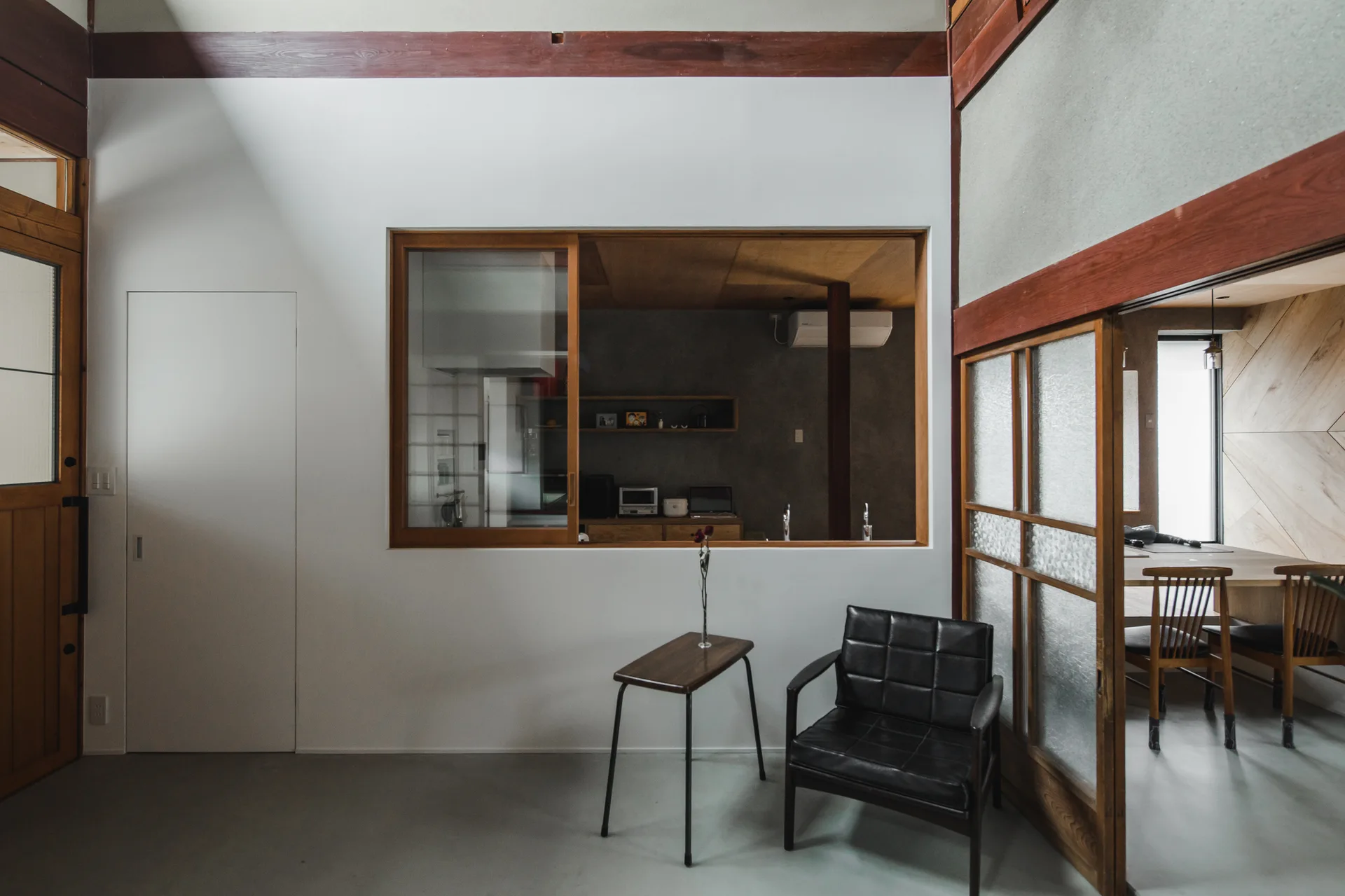 A minimalist living room features a black leather armchair, a slim wood side table, and a large interior window looking into a kitchen.