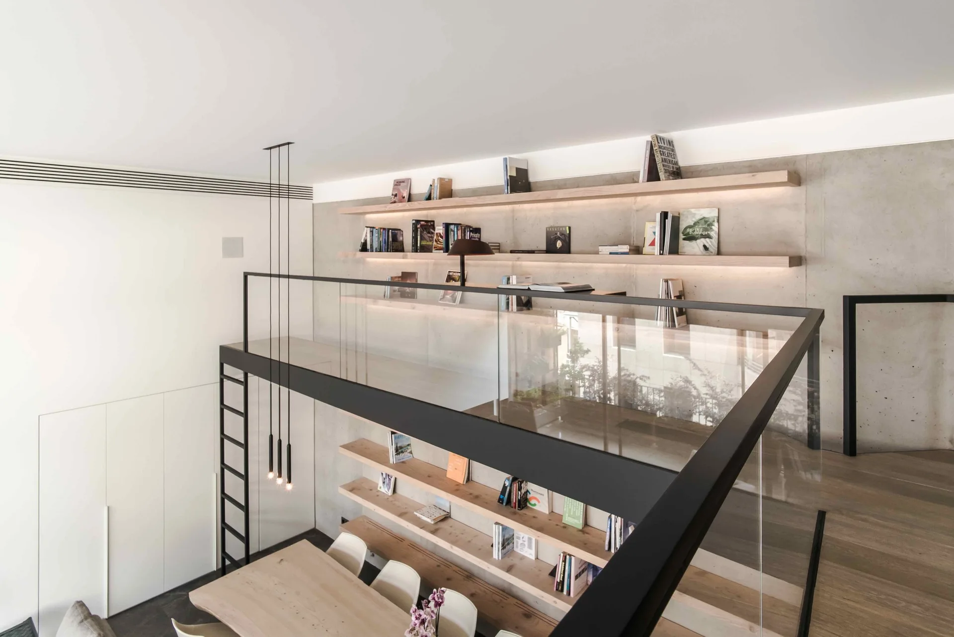 An elevated loft mezzanine features glass railings and long floating wooden bookshelves mounted on a tall exposed concrete wall overlooking a dining table.