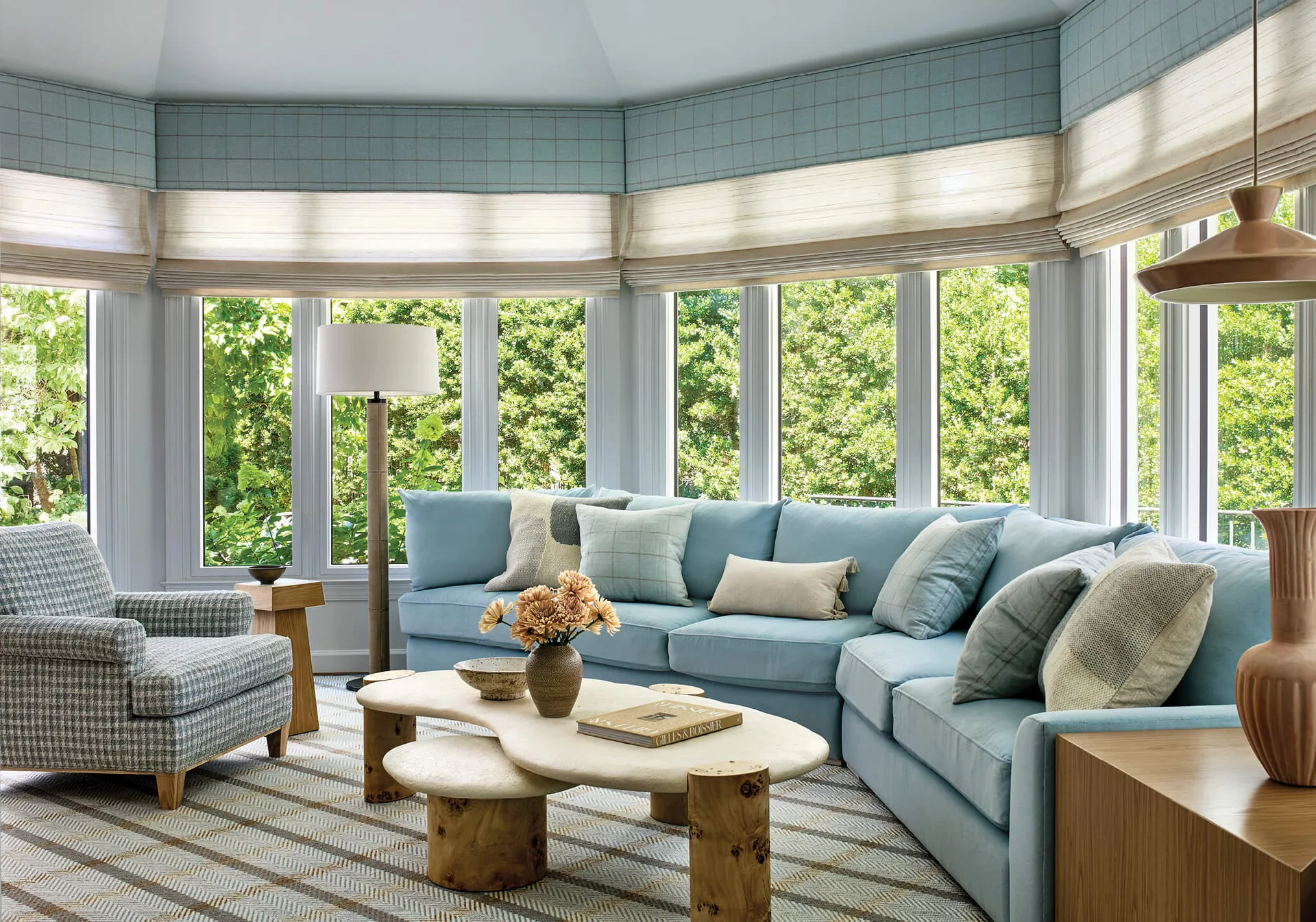 A bright living room featuring a light blue sectional sofa, an organic-shaped stone coffee table, and large bay windows with roman shades.