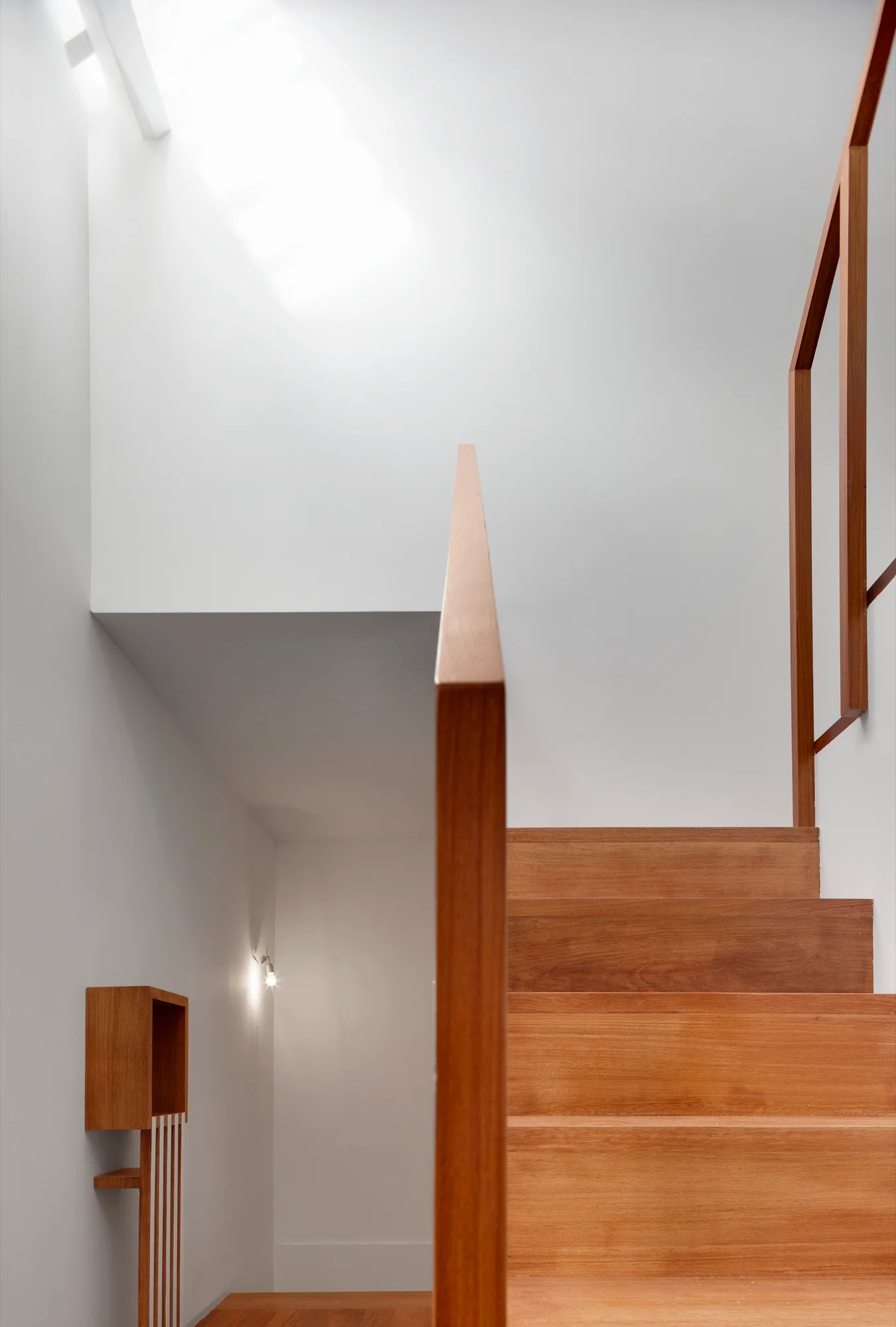 A minimalist wooden staircase with clean timber railings and steps climbs alongside smooth white walls illuminated by an overhead skylight.