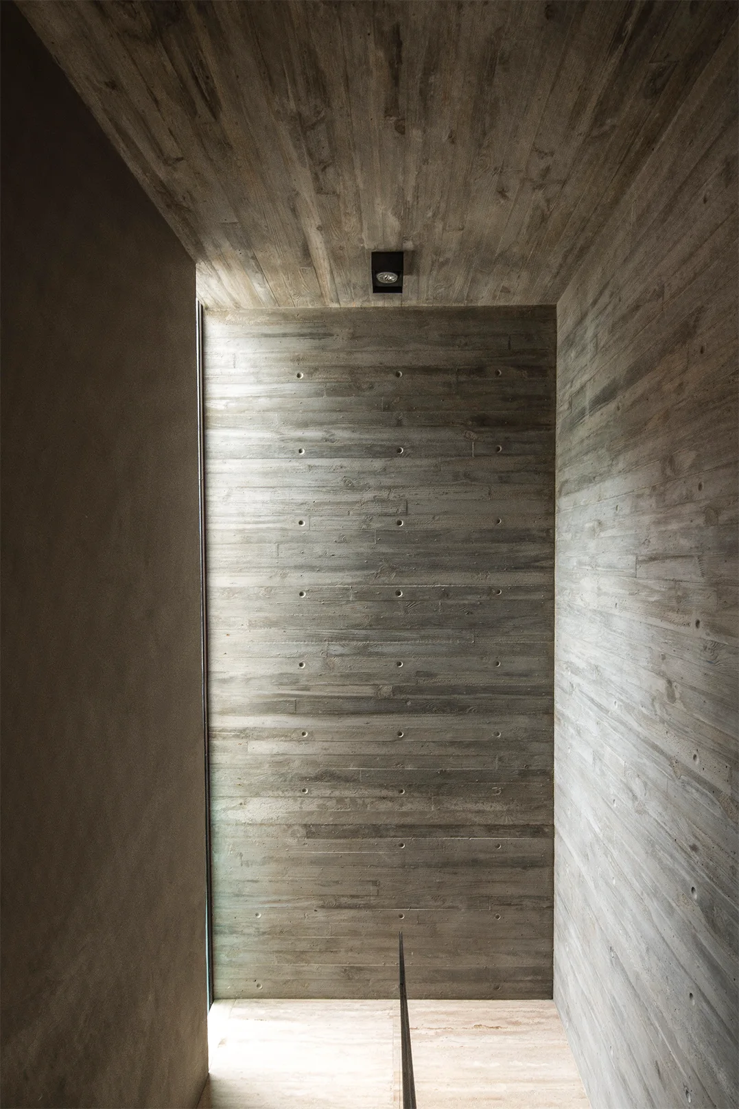 A minimalist stairwell landing features gray board-formed concrete walls, a textured ceiling, and natural light from a slim vertical window.