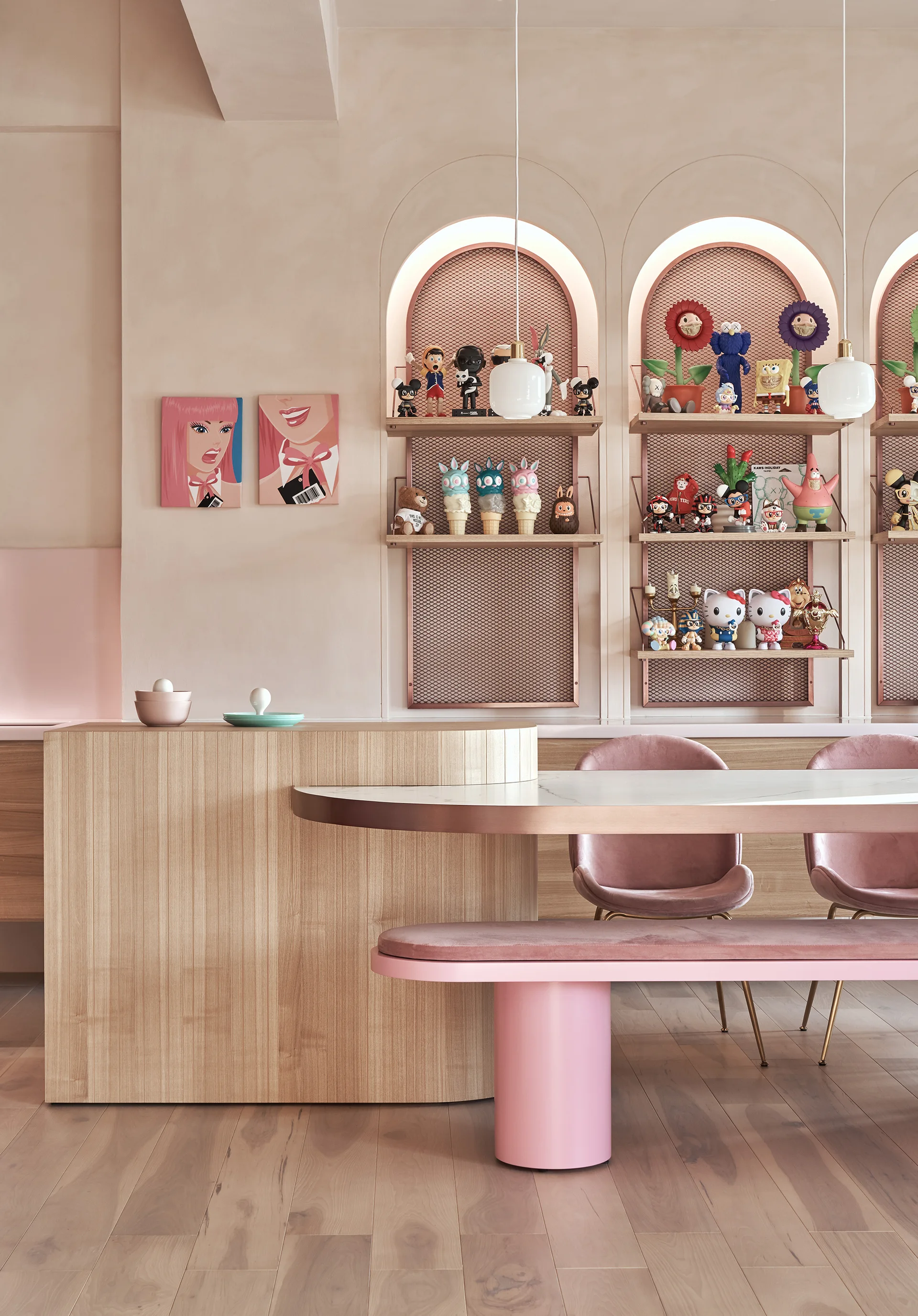 A modern dining room features a light wood table, pink velvet chairs, and a pink bench set against wall-embedded arched display niches with figurines.
