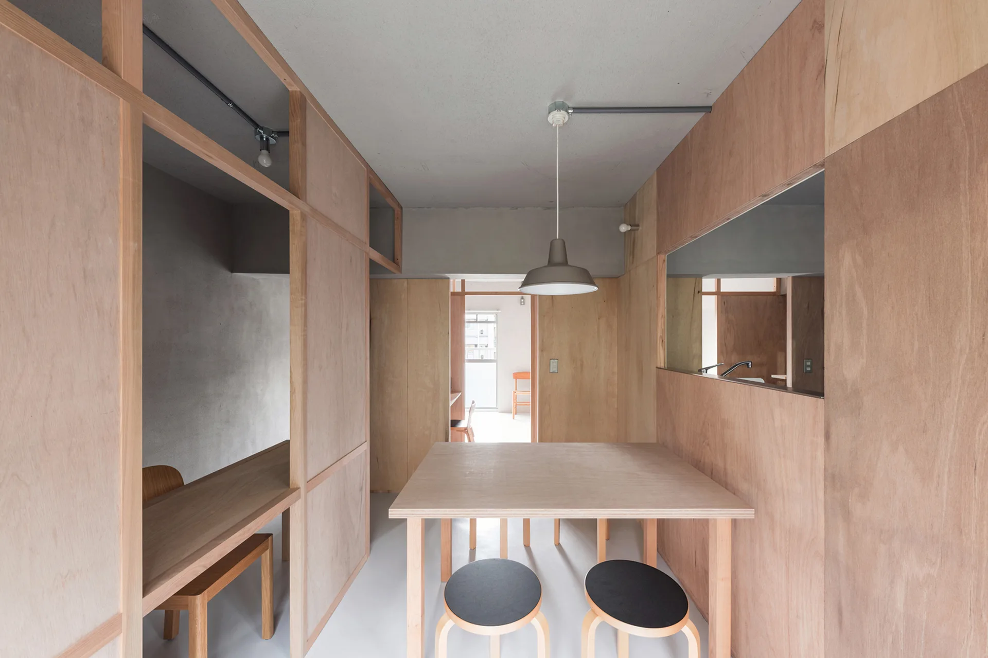 A light wood dining table with two black-topped stools is set against warm plywood walls and a grey ceiling in a minimalist apartment.