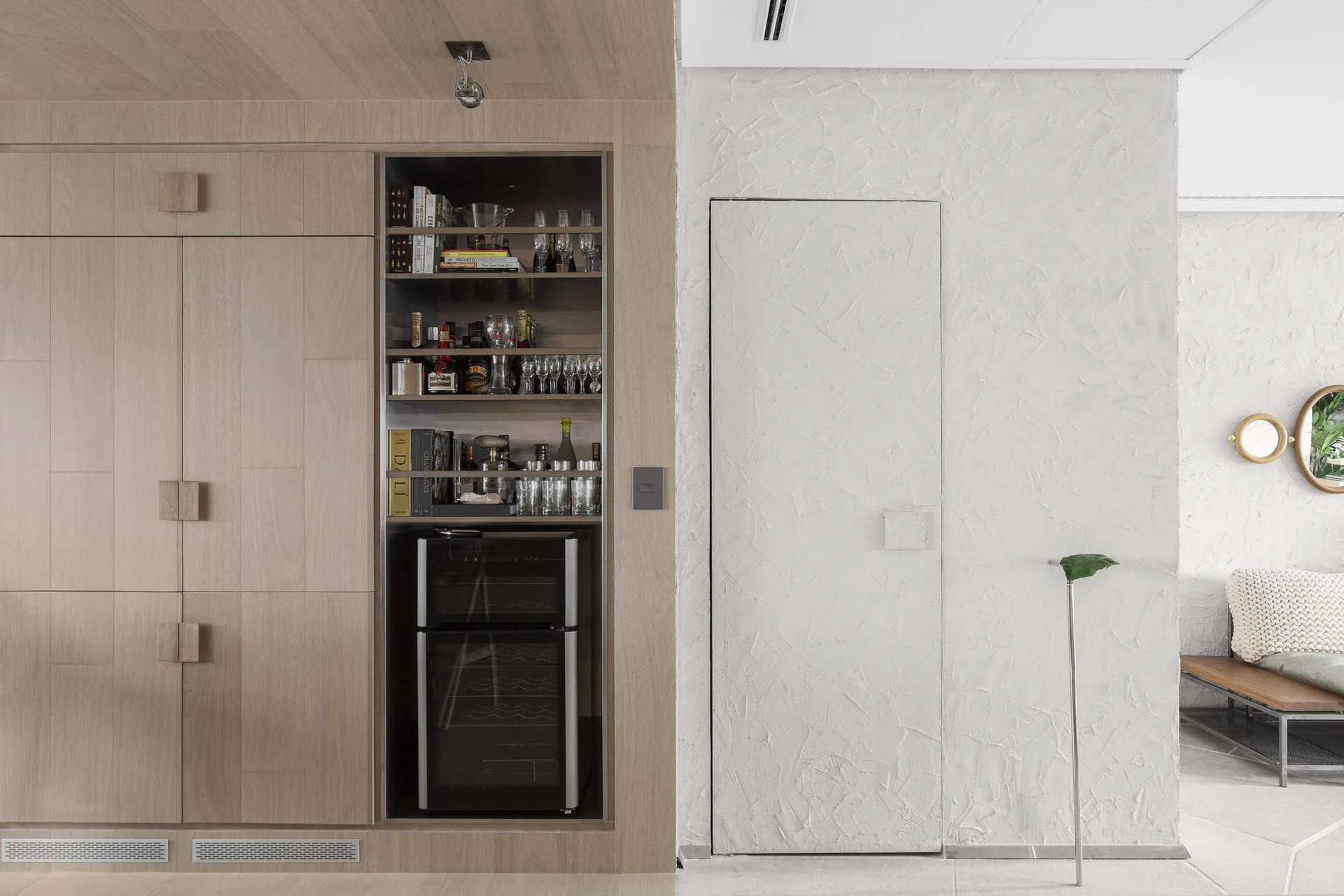A modern home bar features light wood cabinetry with a built-in wine fridge next to a textured plaster wall with a flush-mount hidden door.