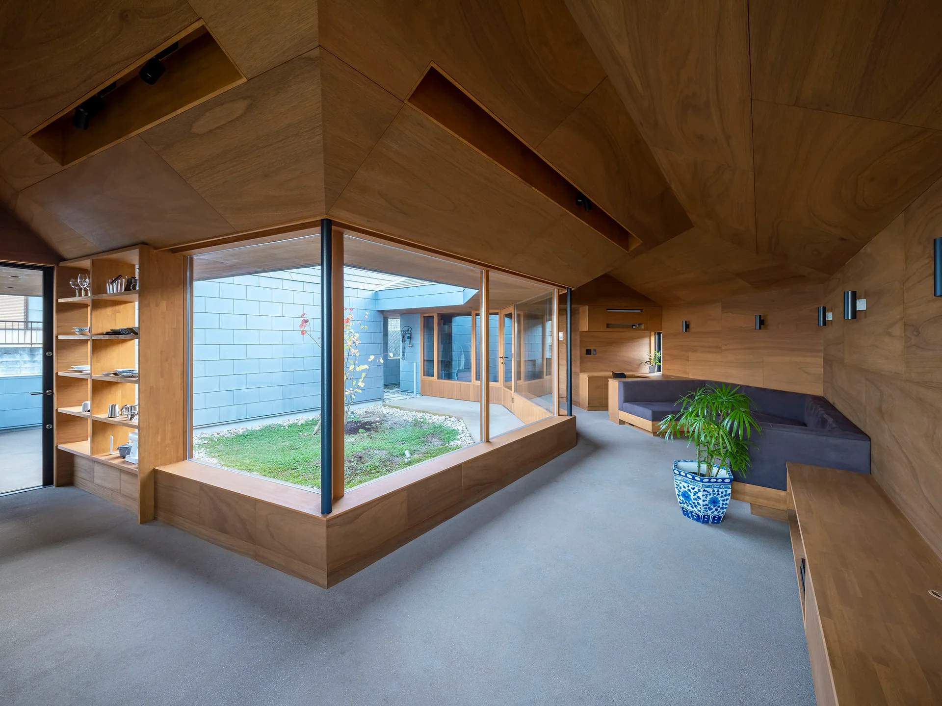 A modern living space featuring wood-paneled walls and ceiling, concrete floors, built-in seating, and a large corner window looking onto an outdoor courtyard.