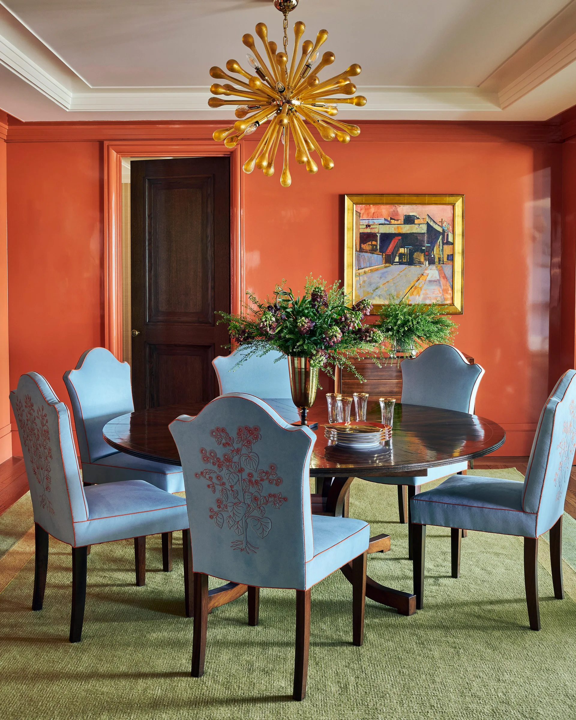 A dining room featuring a round dark wood table, light blue upholstered chairs with coral embroidery, and high-gloss orange walls.