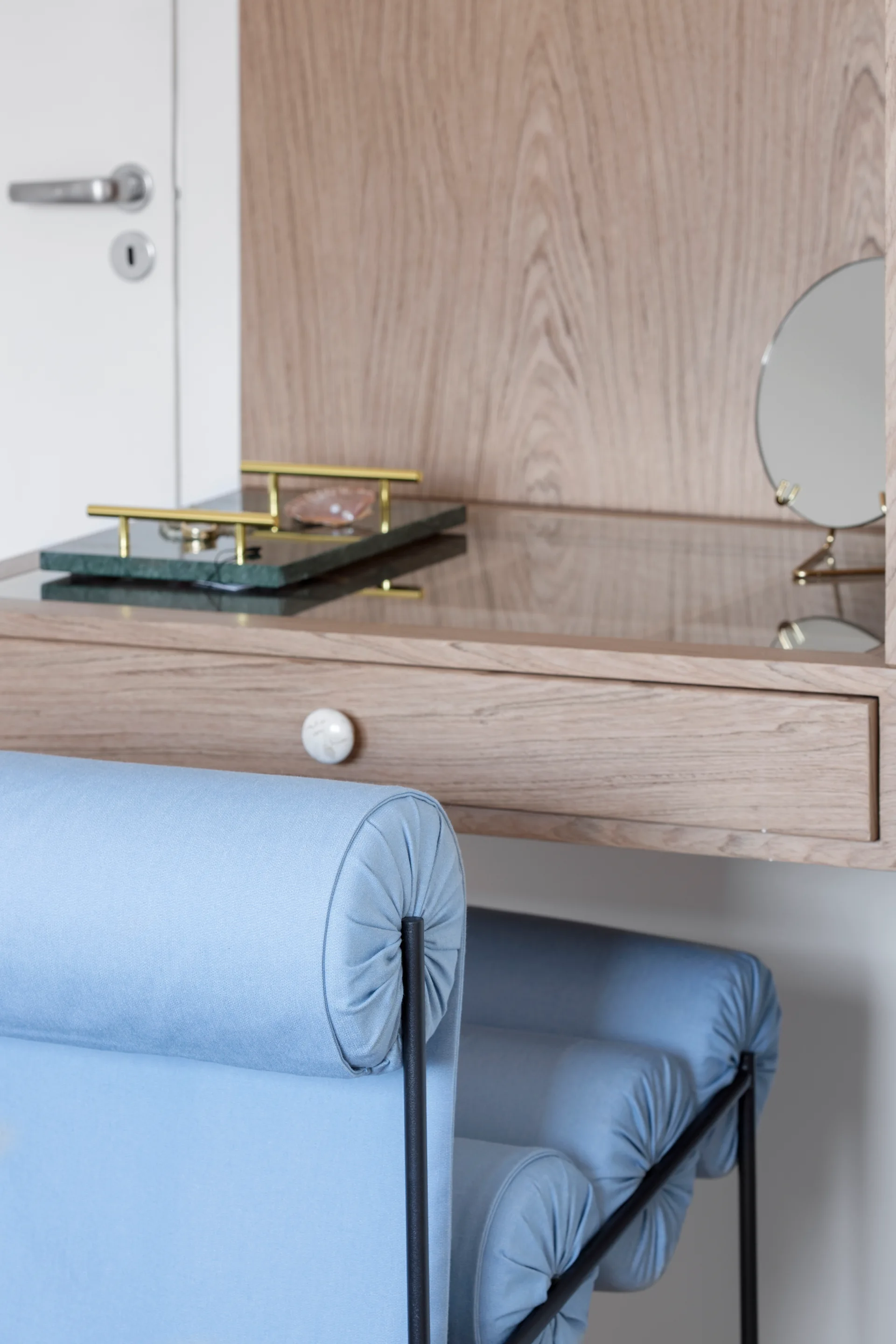 A close-up of a wall-mounted light wood vanity desk paired with a light blue bolster-style stool, a small desktop mirror, and a green marble tray.