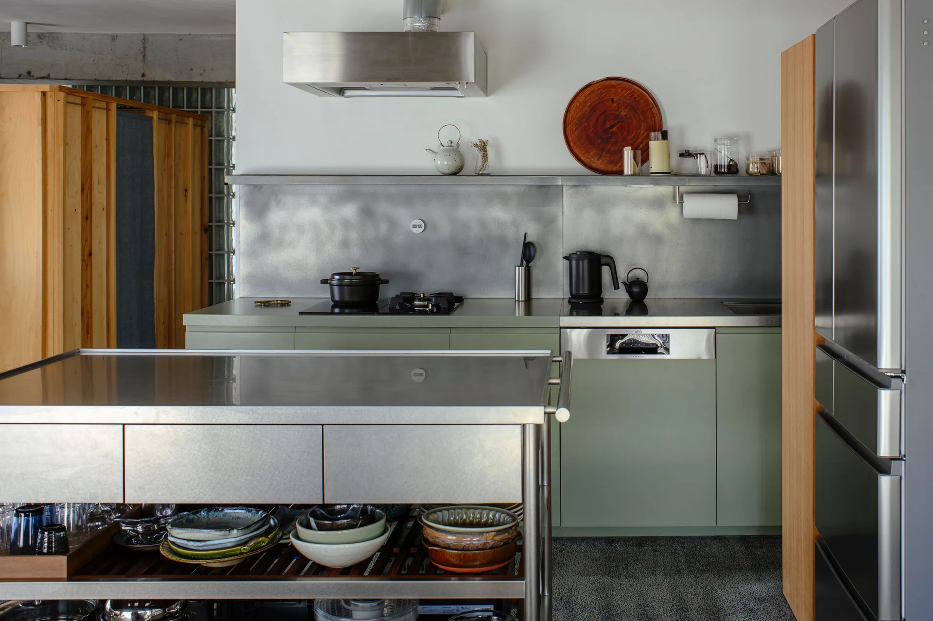 A modern kitchen featuring matte sage green lower cabinets, a brushed stainless steel backsplash, and a metallic prep island with open storage shelving.