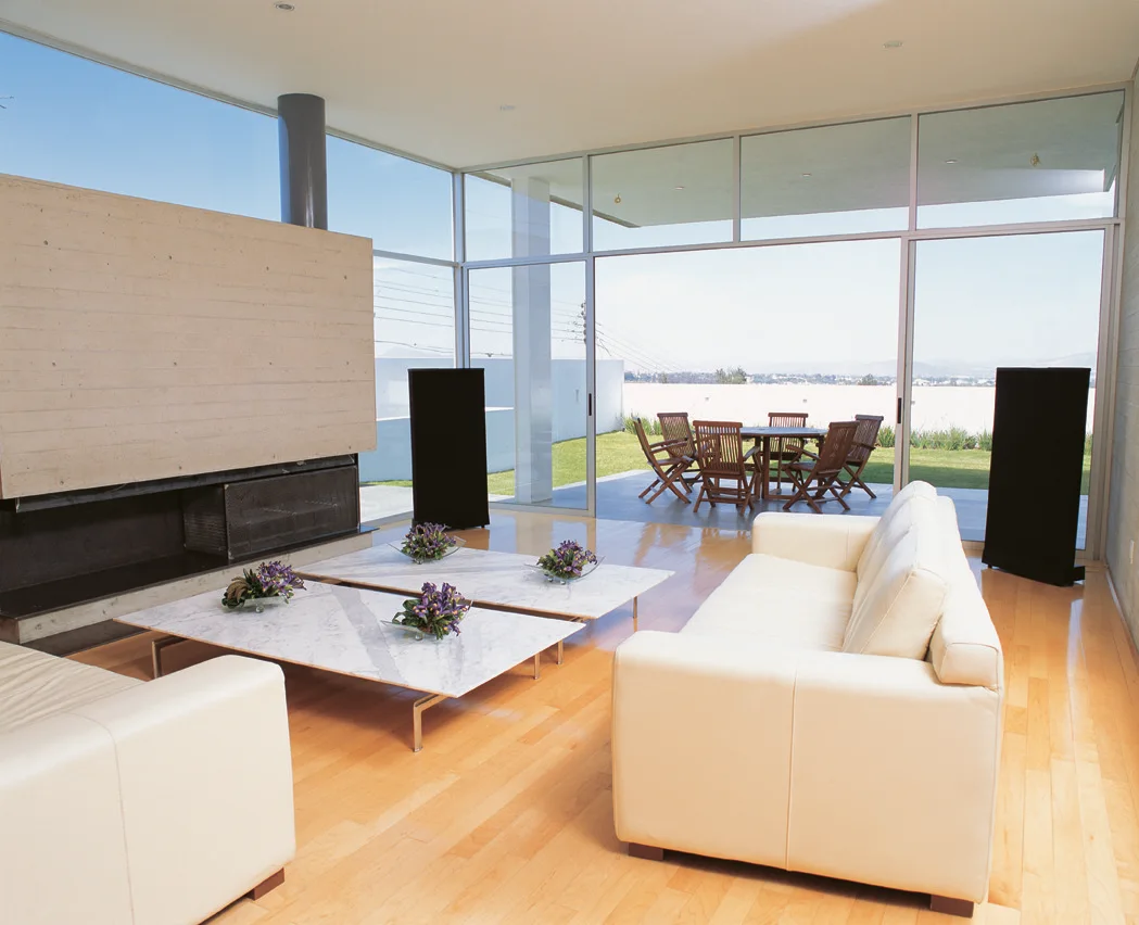 A bright modern living room with two white leather sofas, low marble coffee tables, and large glass walls opening to an outdoor patio.