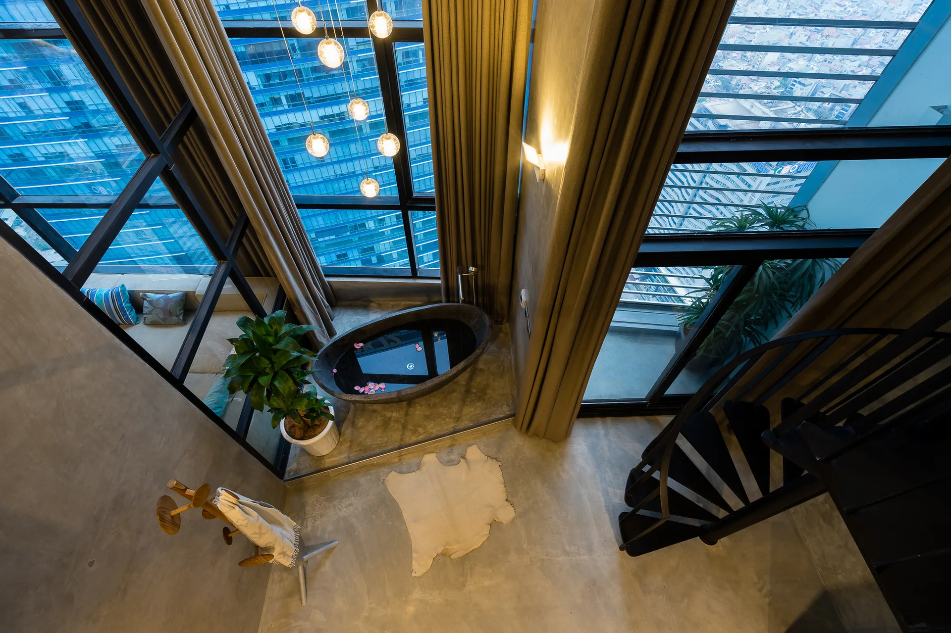 High-angle view of a modern loft bathroom featuring a black stone bathtub, concrete floor, spiral staircase, and large floor-to-ceiling windows.