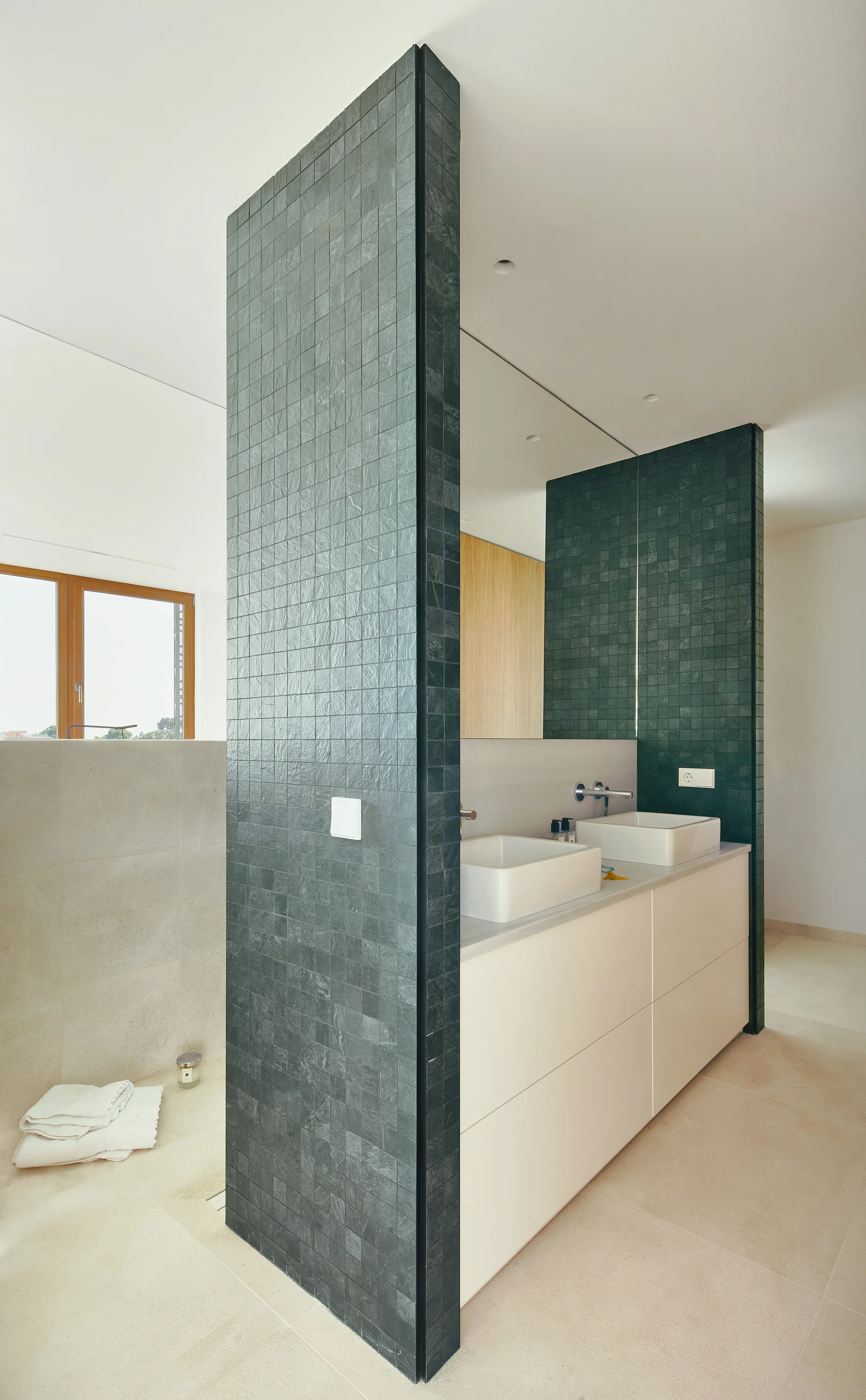 A modern bathroom features a white double vanity and mirror set against a floor-to-ceiling partition covered in small dark green tiles.