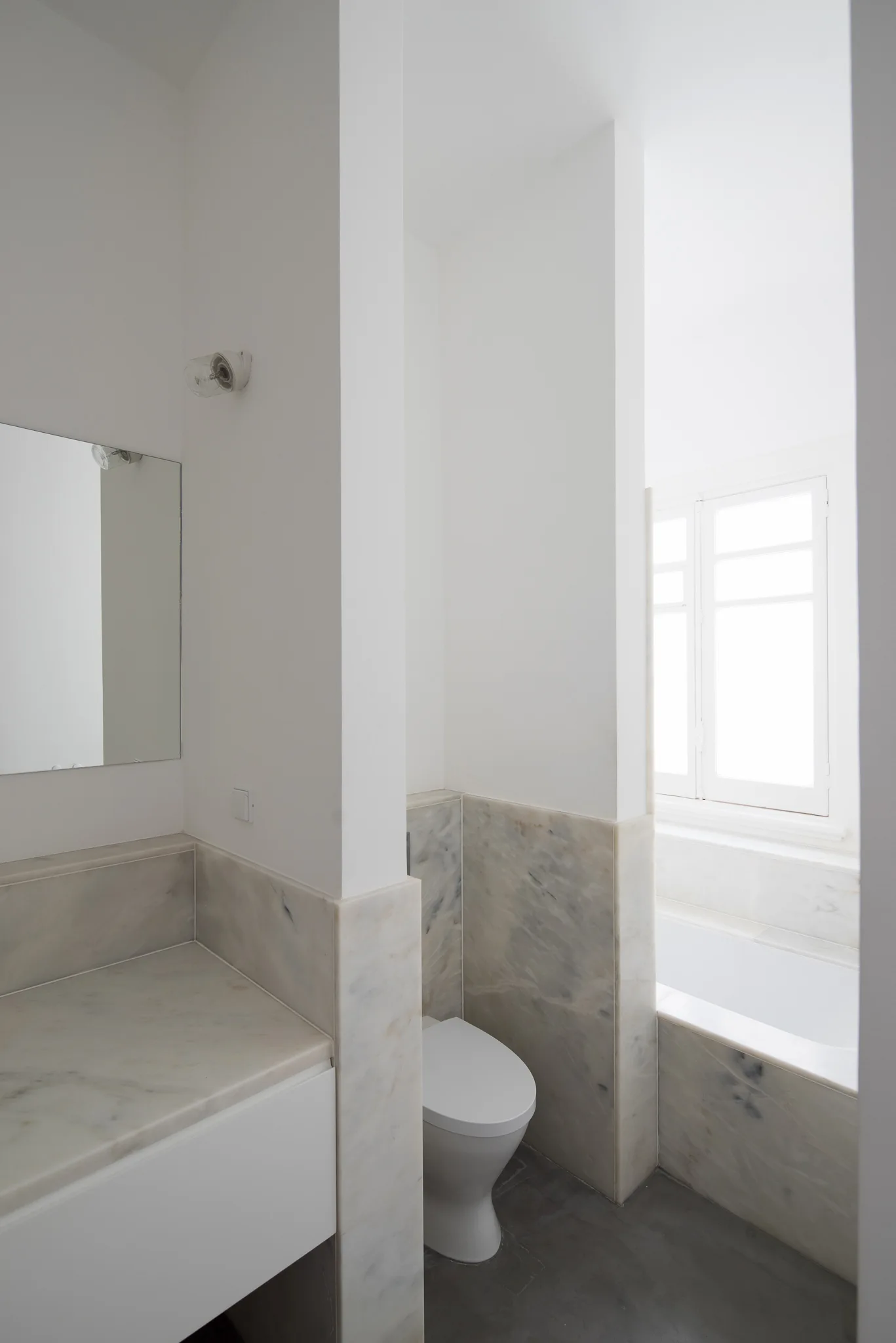 A minimalist white bathroom features low marble wall cladding, a built-in bathtub under a window, and partitioned vanity and toilet areas.