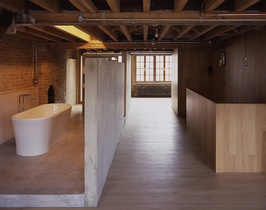 An industrial loft bathroom featuring a white freestanding bathtub positioned next to a raw concrete partition wall beneath exposed wooden ceiling beams.