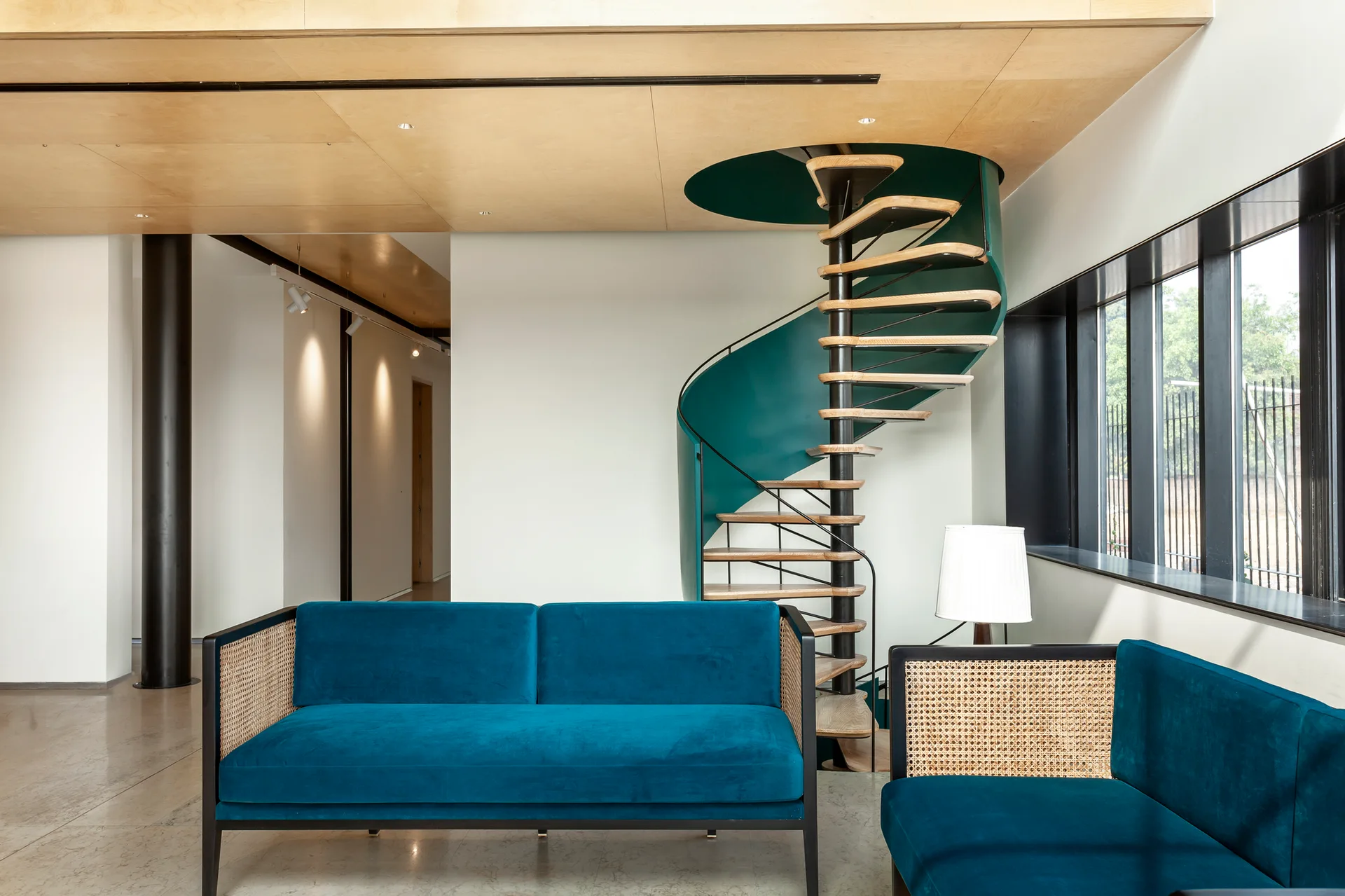 A modern living room features a blue velvet sofa with cane sides, a striking teal spiral staircase, and a wood-paneled ceiling.