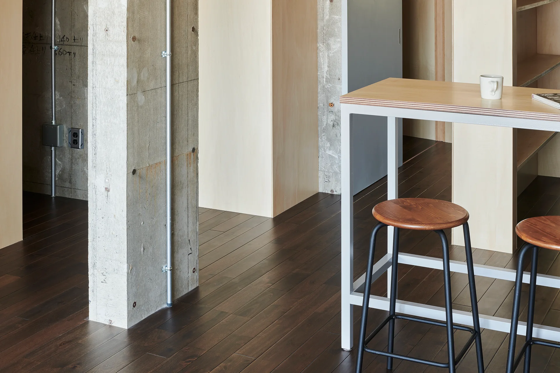 An industrial room features a dark wood floor, a raw concrete column with metal conduits, and a high plywood table with two round stools.