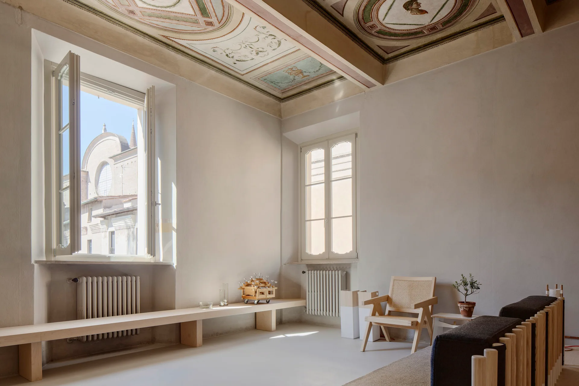 A minimalist living room features a long light wood bench below windows, a cane armchair, and an ornate frescoed ceiling.