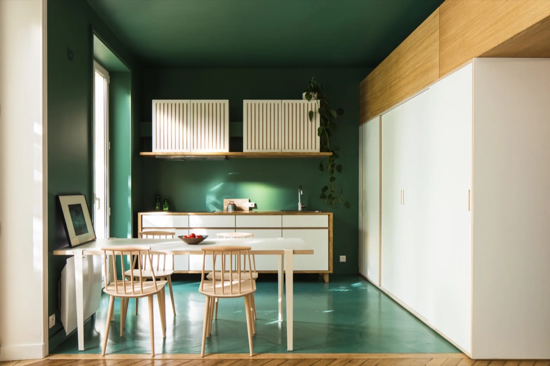 A modern kitchen featuring forest green walls, ceiling, and floor, balanced by white cabinetry, a white dining table, and light wood spindle-back chairs.