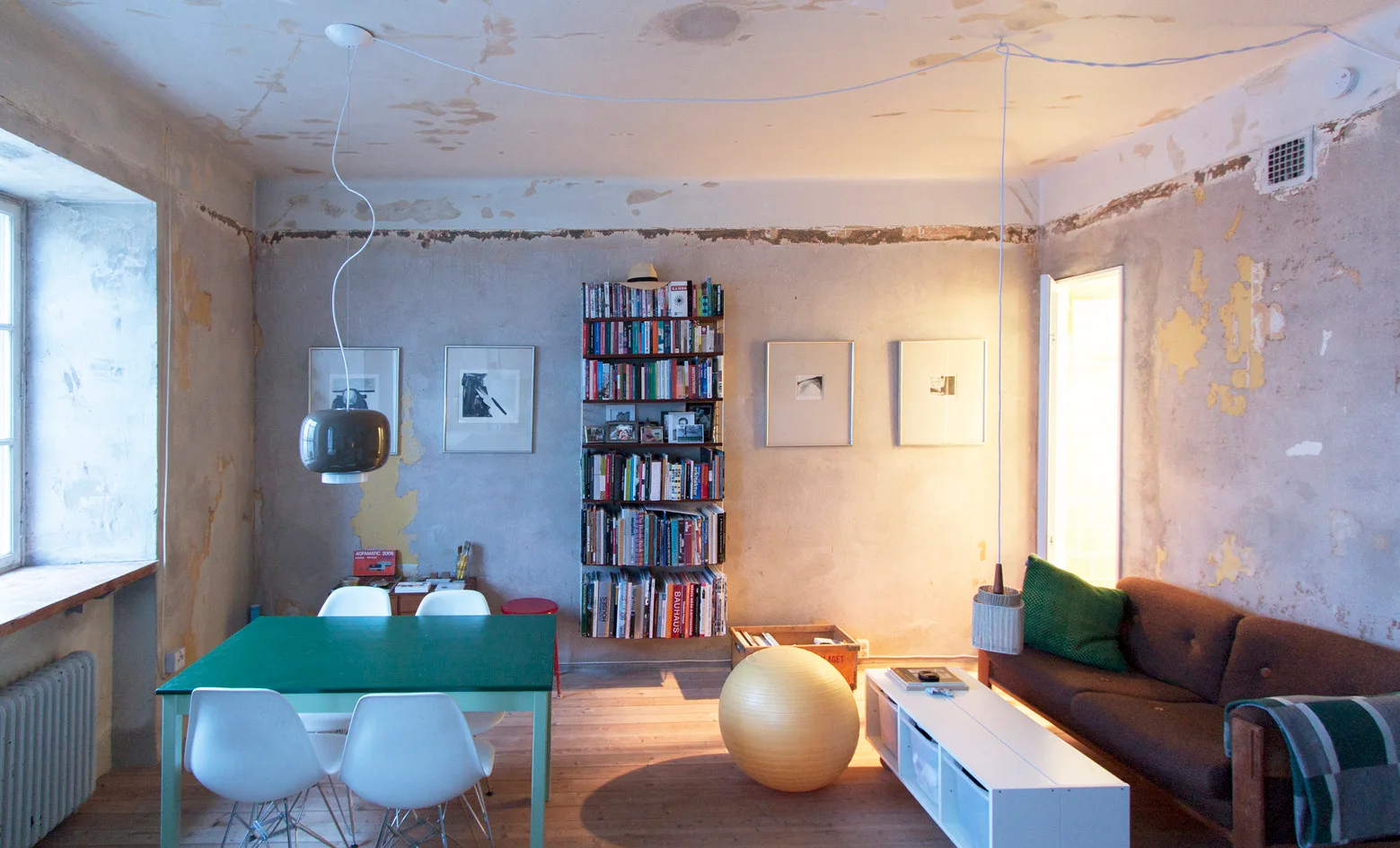 An open dining and living space features textured, unfinished plaster walls, a bright green dining table with white chairs, and a brown fabric sofa.