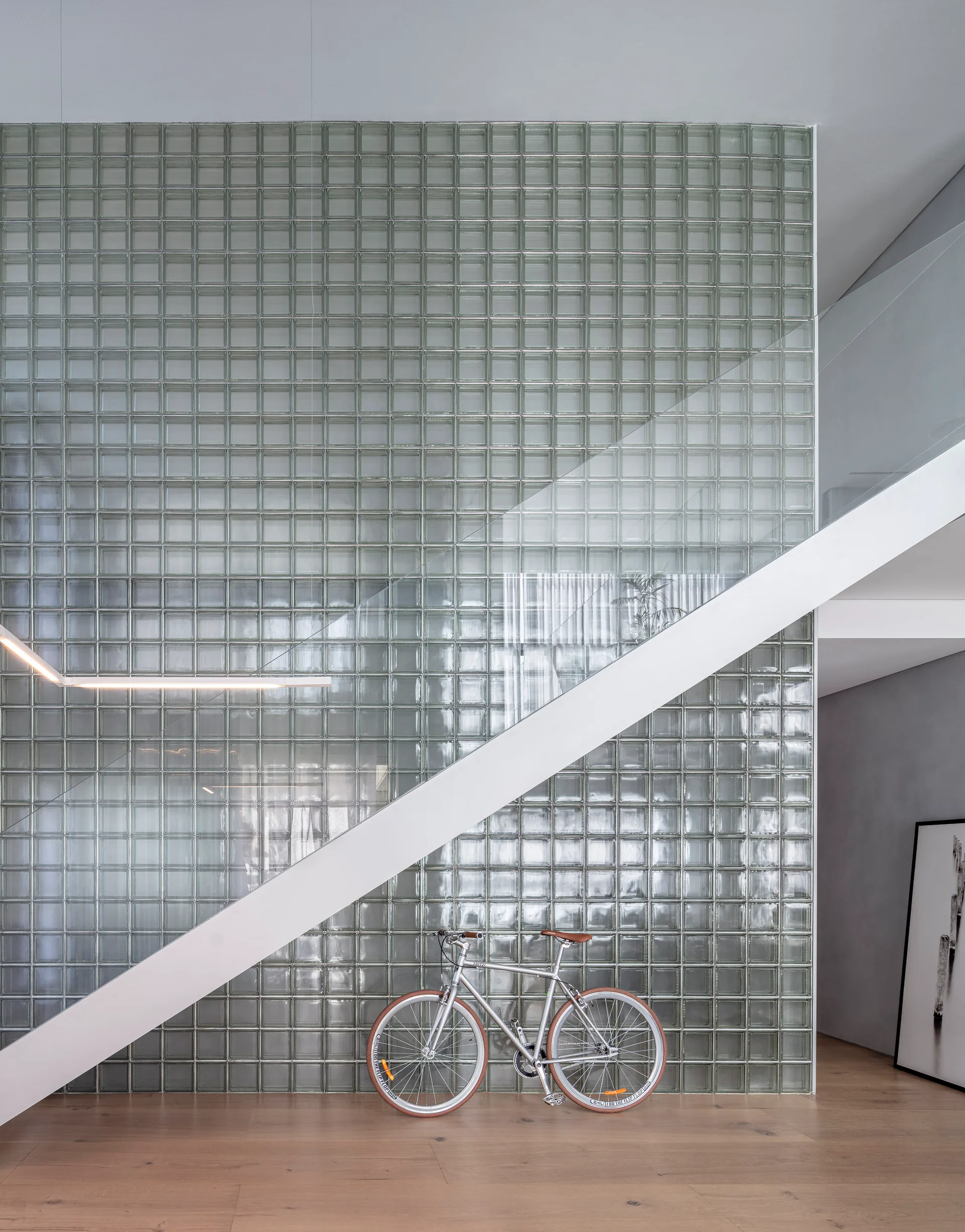 A white structural staircase with a glass railing ascends in front of a floor-to-ceiling translucent glass block wall on light wood floors.
