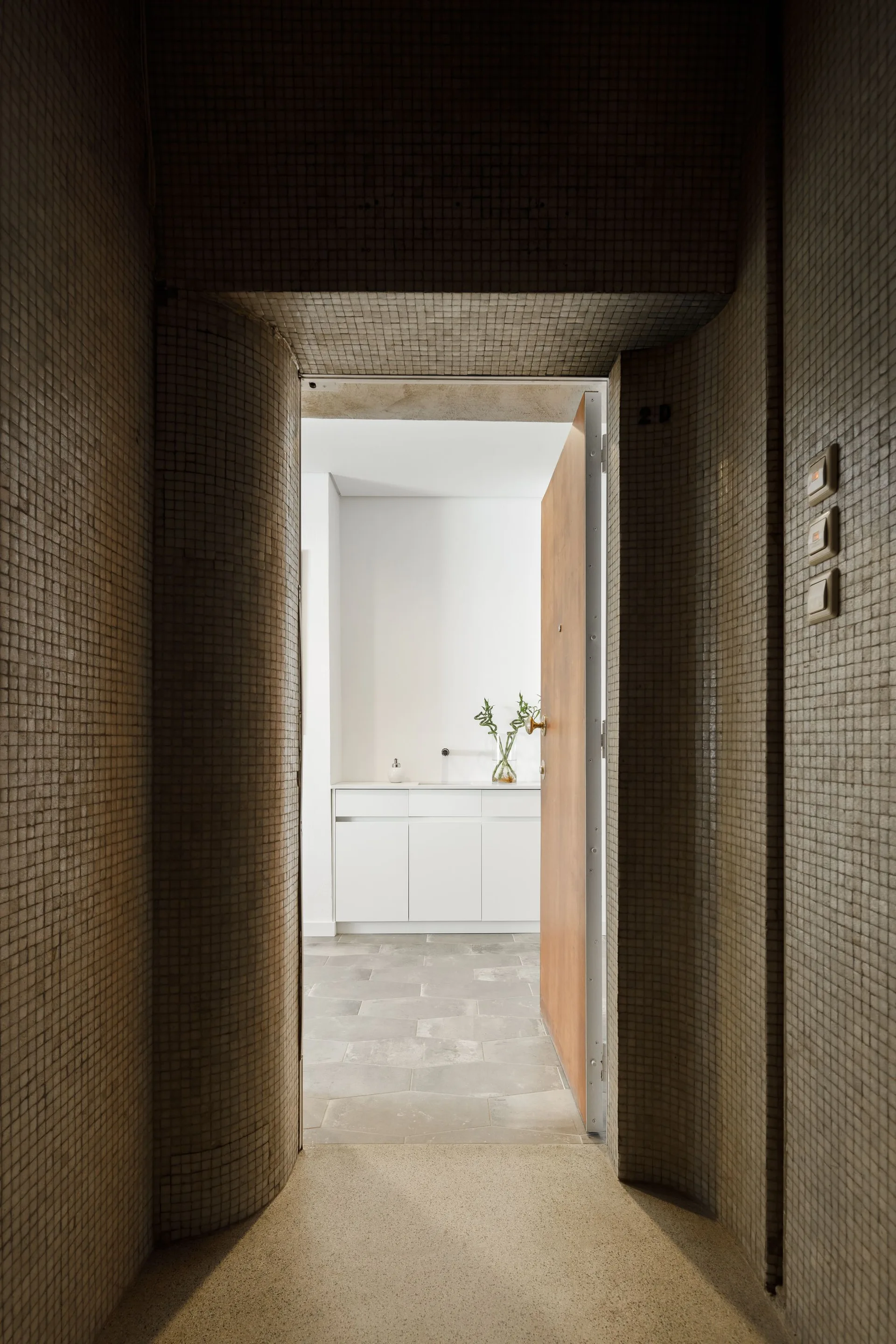 A narrow arched corridor covered in small beige mosaic tiles leads to an open wooden door and a bright bathroom with a white vanity.