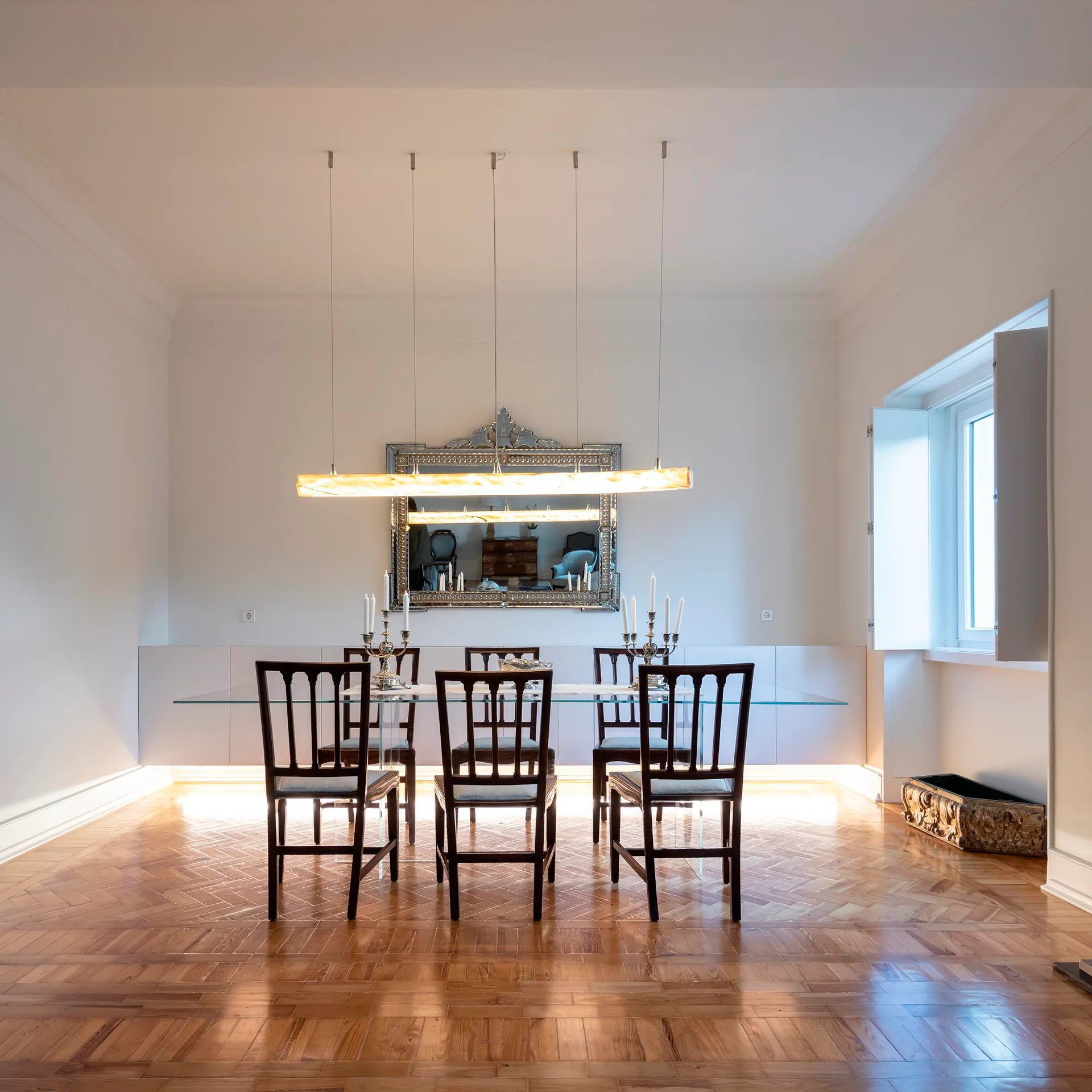 A formal dining room features a glass-topped dining table with dark wood chairs, herringbone wood floors, a large mirror, and a linear pendant light.
