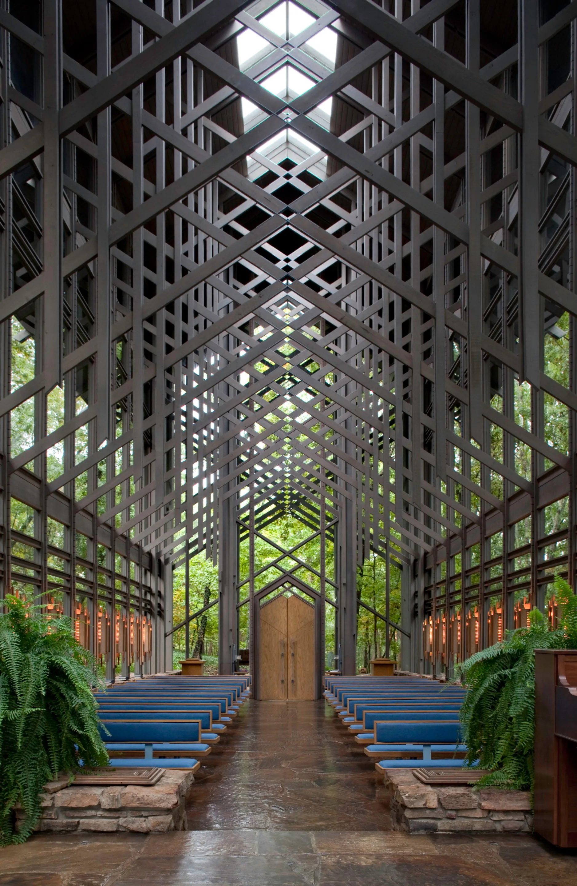 A symmetrical chapel interior features intricate intersecting dark wood roof trusses, glass walls showing forest greenery, and rows of blue-cushioned pews.
