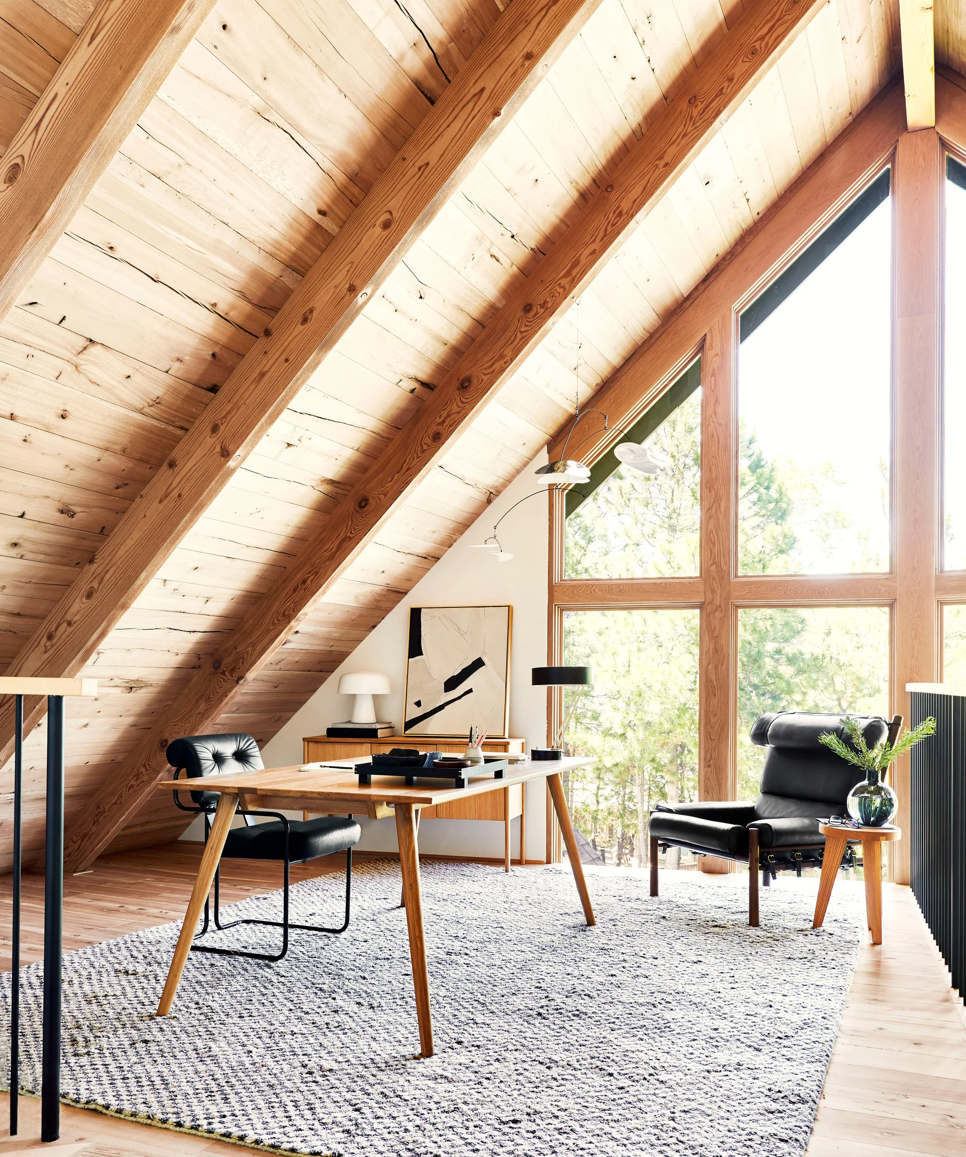 A lofted home office features a wooden desk, black office chairs, and a textured area rug under a high sloped wood plank ceiling.