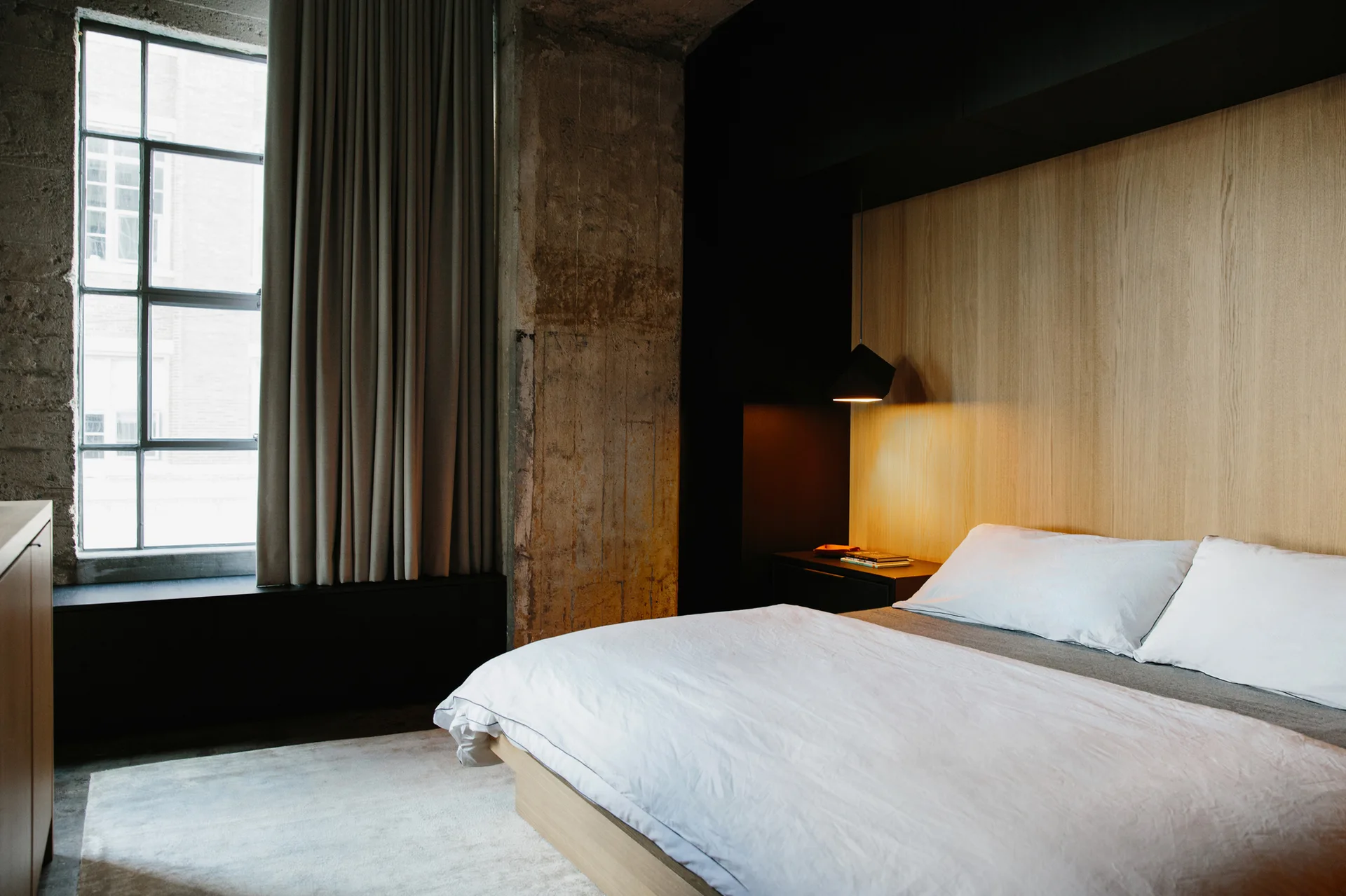 A minimalist platform bed rests against a light wood accent wall next to a large grid window and raw concrete pillar.