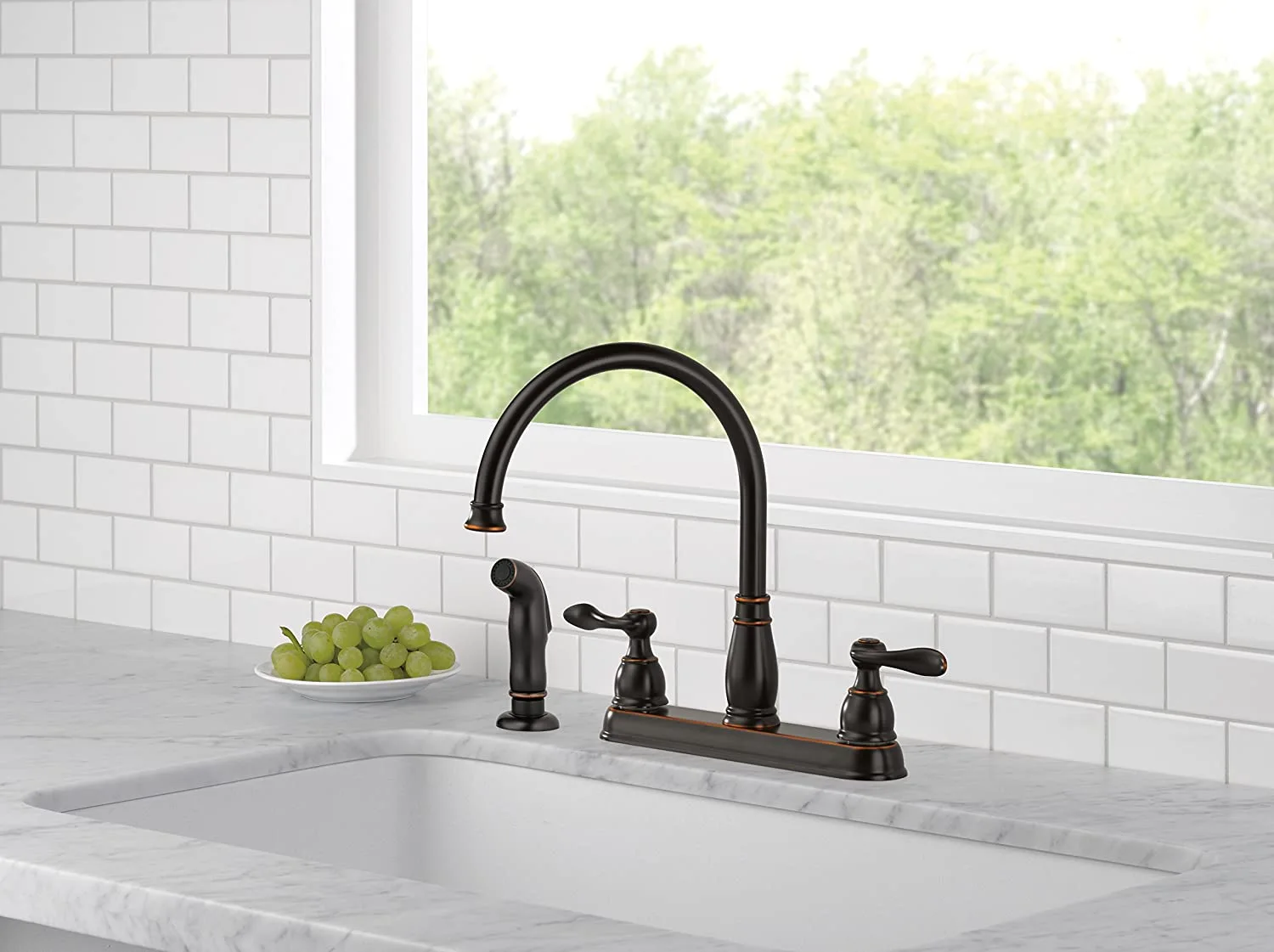 An oil-rubbed bronze faucet and side sprayer are installed on a marble countertop with a white undermount sink and subway tile backsplash.