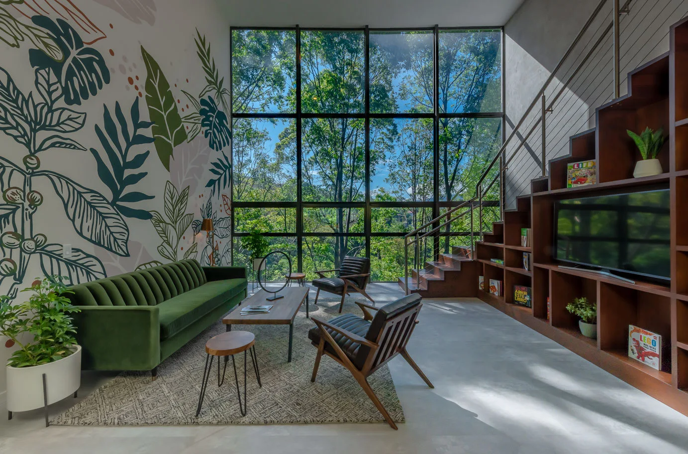 A modern living room features a green velvet sofa, a botanical wall mural, and a large grid window overlooking a forest.