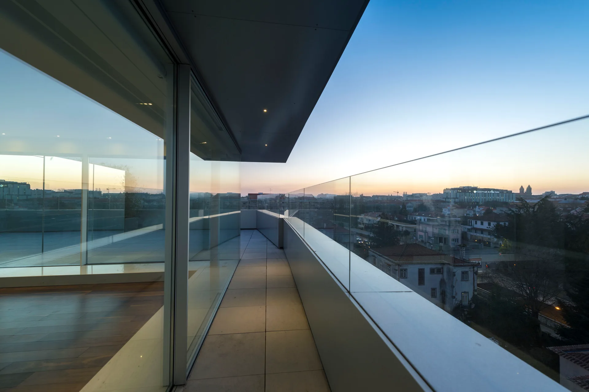 A minimalist outdoor balcony features a frameless glass railing, large grey stone floor tiles, and a floor-to-ceiling glass wall at sunset.