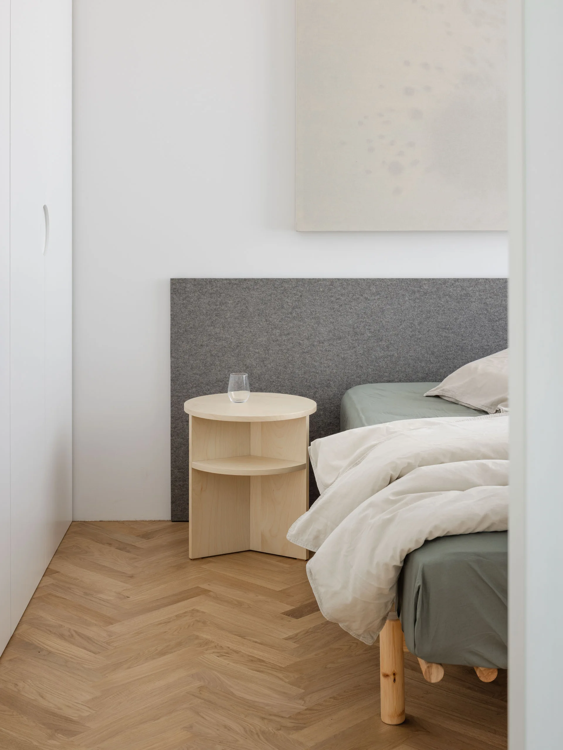 A minimalist bedroom features a grey felt headboard behind a bed with sage green linen, a circular wood nightstand, and herringbone hardwood flooring.