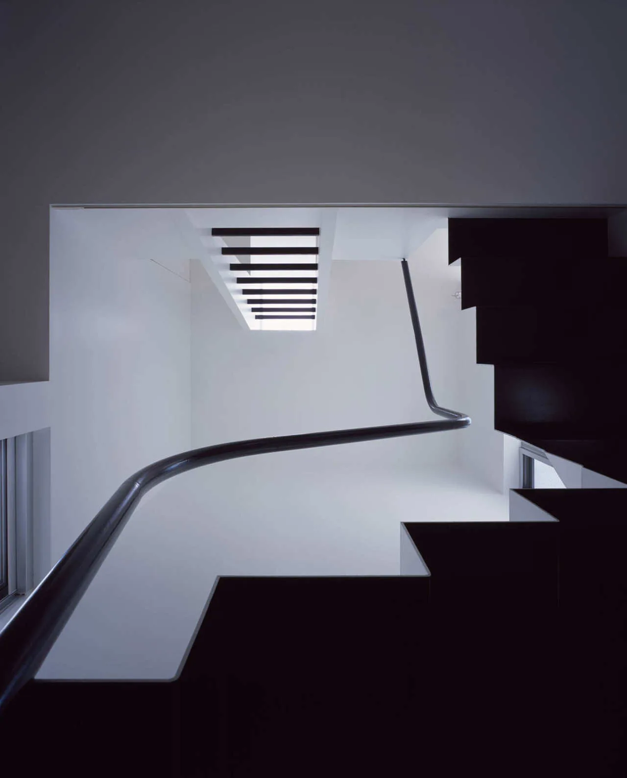 A black minimalist staircase and curved metal handrail curve upward through a stark white stairwell under a rectangular skylight.