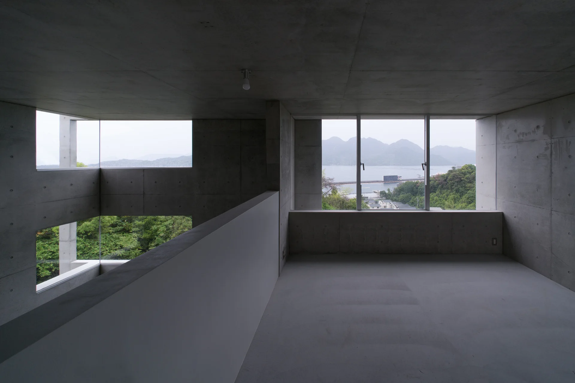 An empty minimalist concrete room features large rectangular windows overlooking a green hillside, a body of water, and mountains.