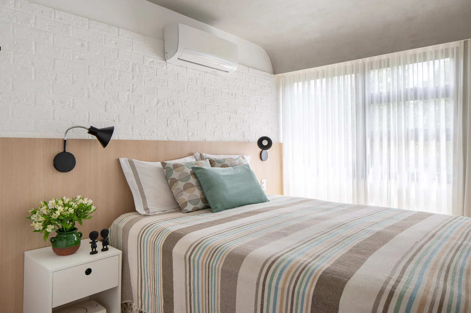 A minimalist bedroom features a striped duvet, a light wood paneled headboard against a white brick wall, and a bedside table with a green vase.