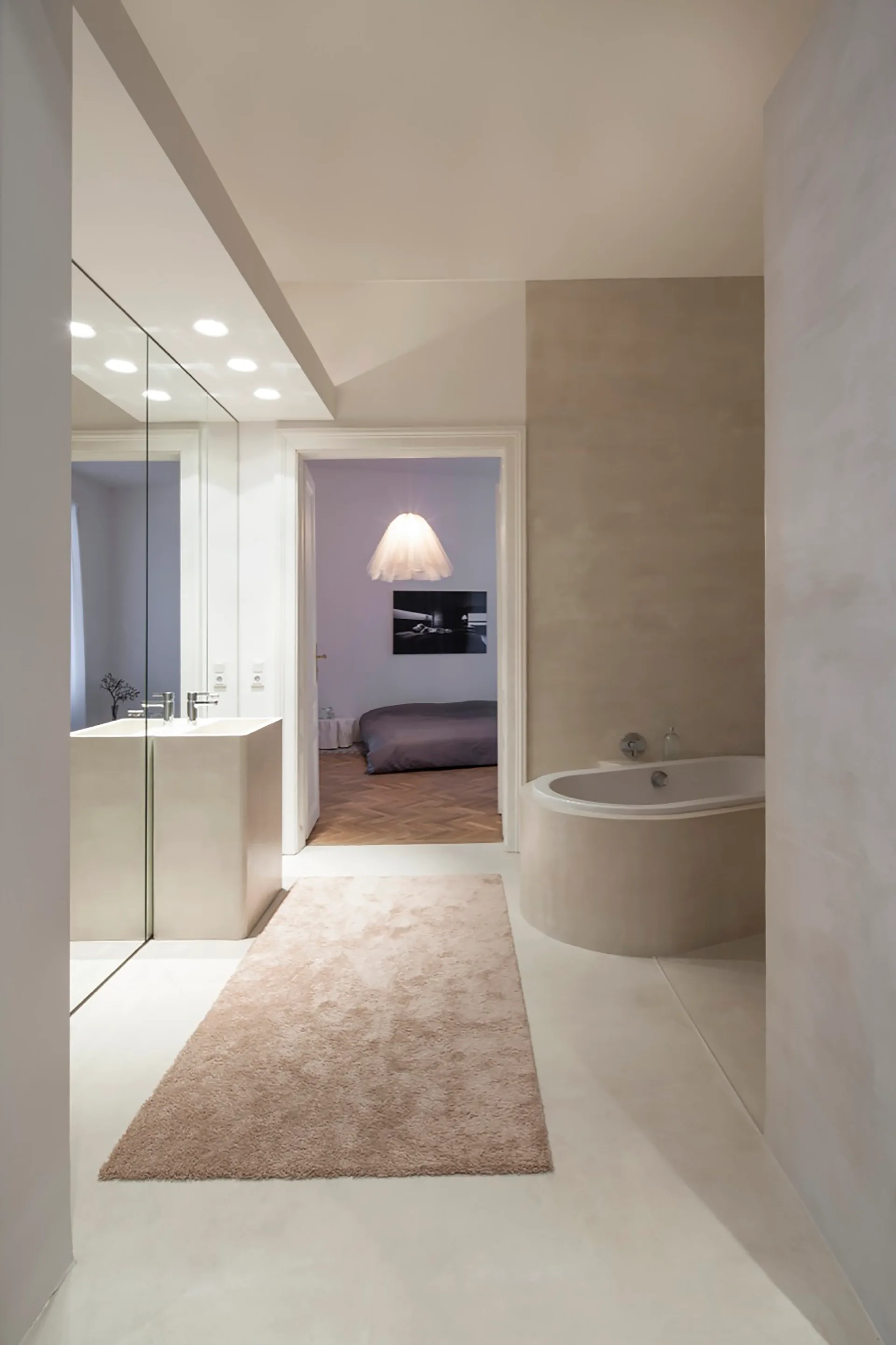 A minimalist beige bathroom features a monolithic plaster sink, a built-in oval bathtub, and an open doorway looking into an adjoining bedroom.