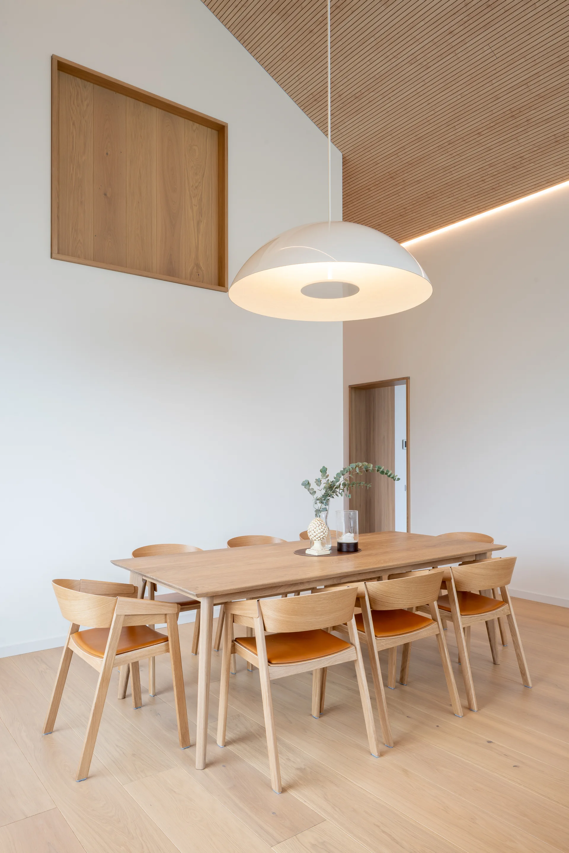 A light oak dining set with orange leather cushions sits beneath a large white dome pendant light and a slatted wood ceiling.