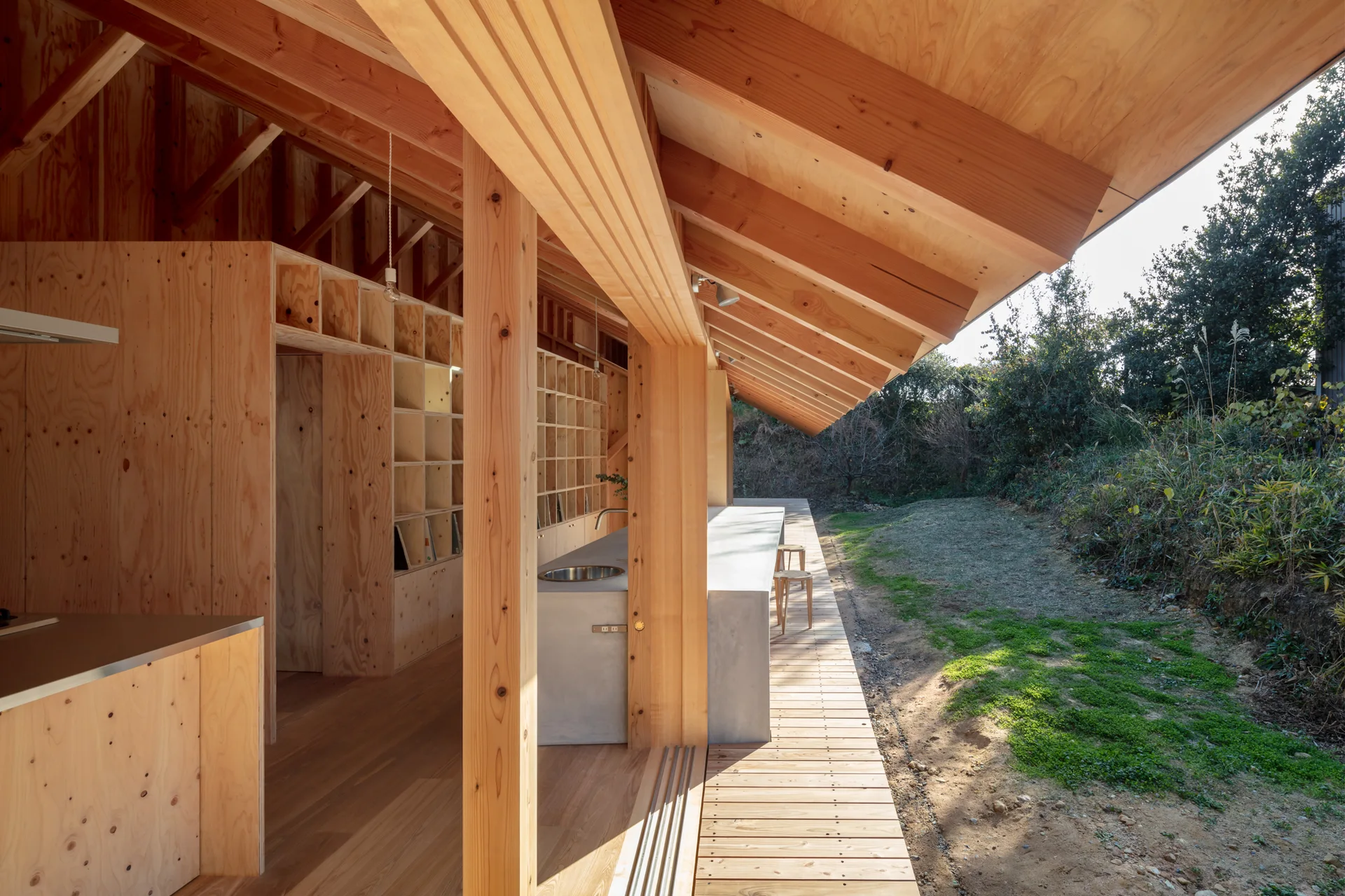 A minimalist kitchen features natural plywood walls, a concrete island, and exposed timber ceiling beams opening to an outdoor wooden deck.
