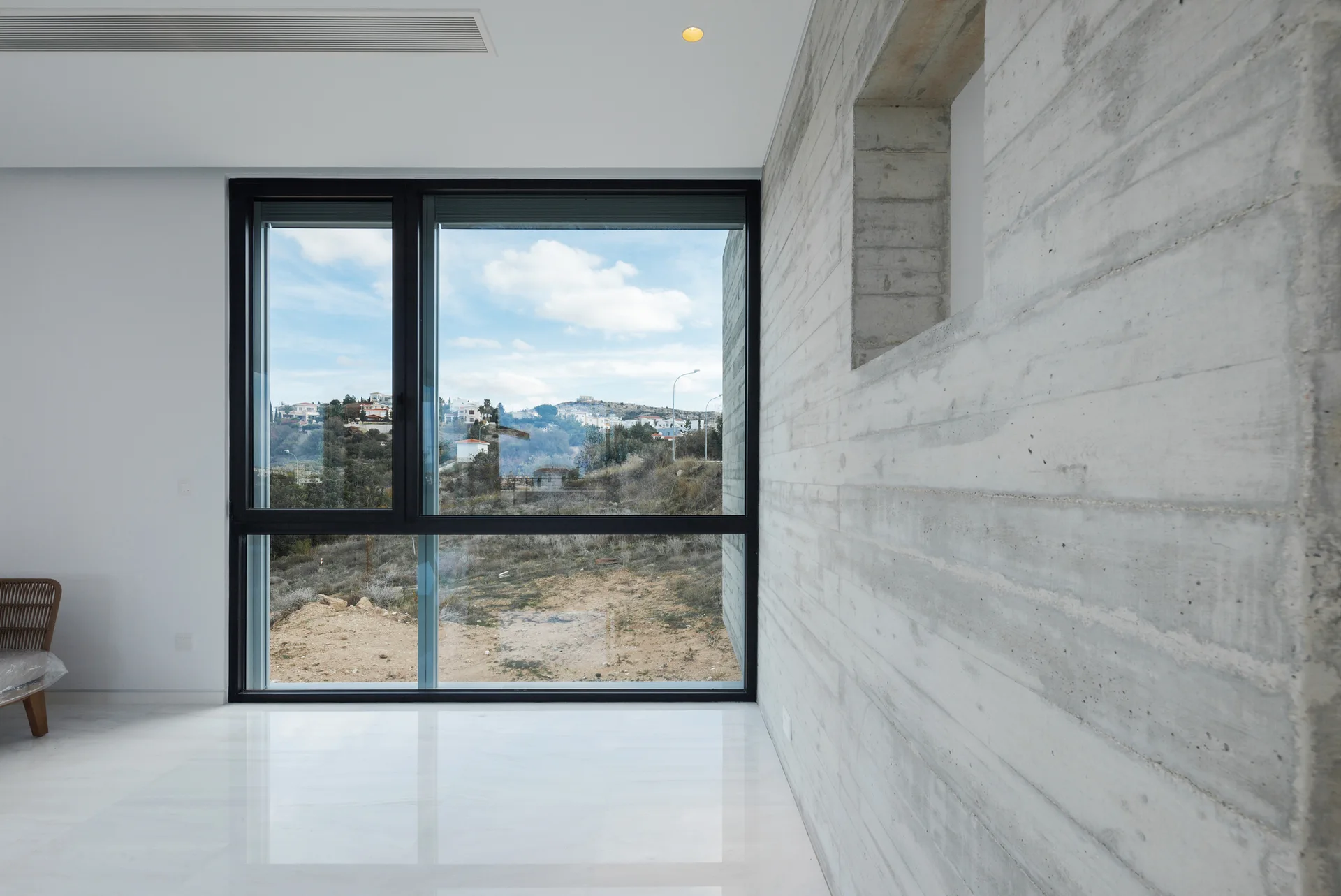 A minimalist room features a textured board-formed concrete wall, highly reflective white flooring, and a large window looking out onto a landscape.