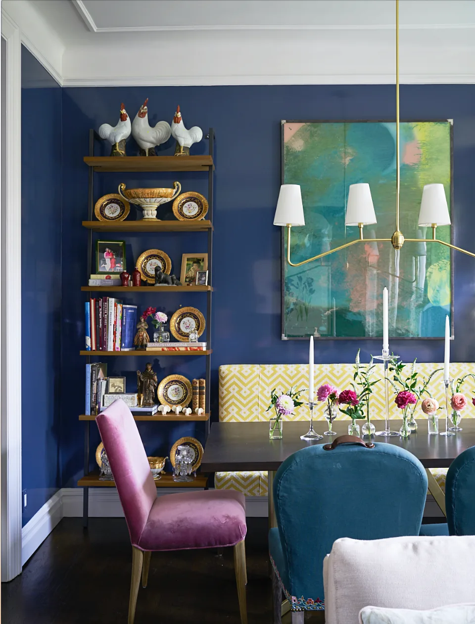 A dining room features high-gloss navy walls, a yellow chevron bench, velvet chairs, a dark wood table, and an open shelf displaying plates and books.