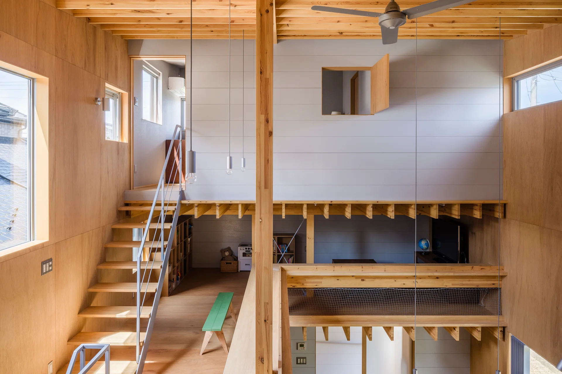 A multi-level minimalist home interior featuring light plywood wall panels, an open-riser wooden staircase with grey metal railing, and exposed ceiling beams.