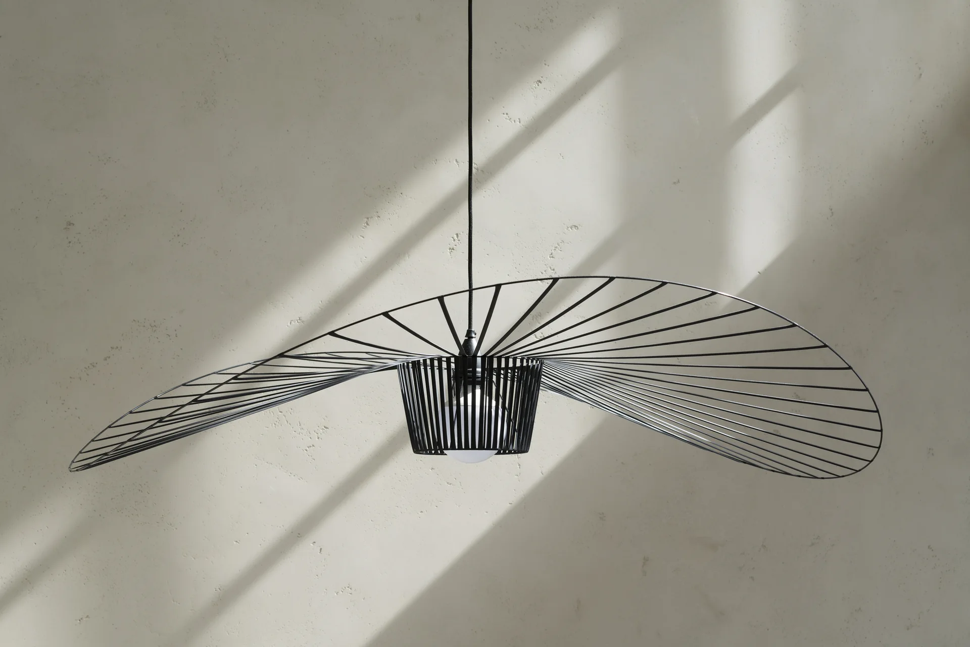 A black metal wire-frame pendant light with a wide undulating brim hangs against a textured beige plaster wall.