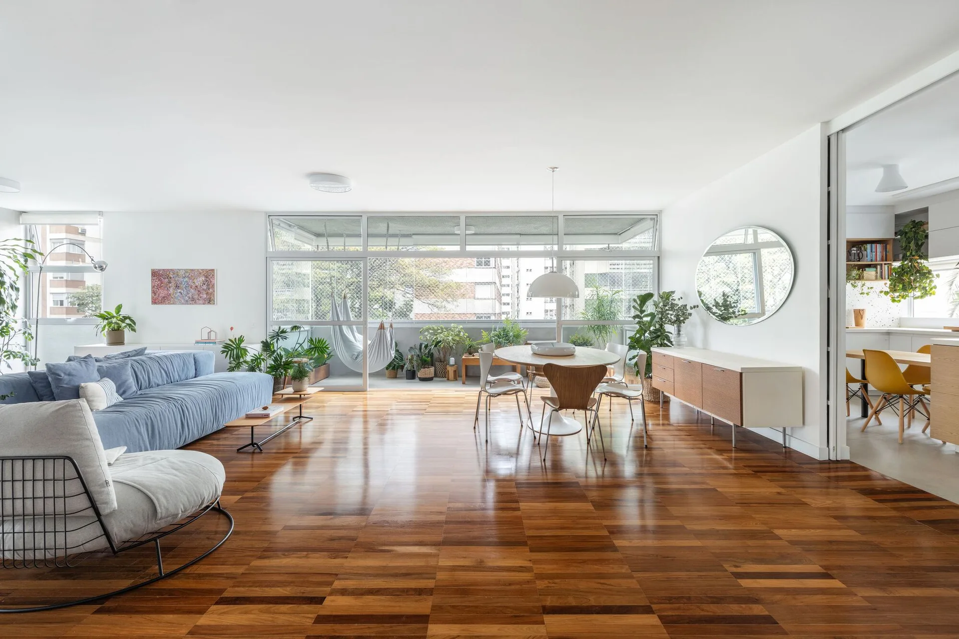 A bright open-plan apartment interior with checkered hardwood floors, a blue sofa, a round dining table, and large glass doors opening to a balcony.