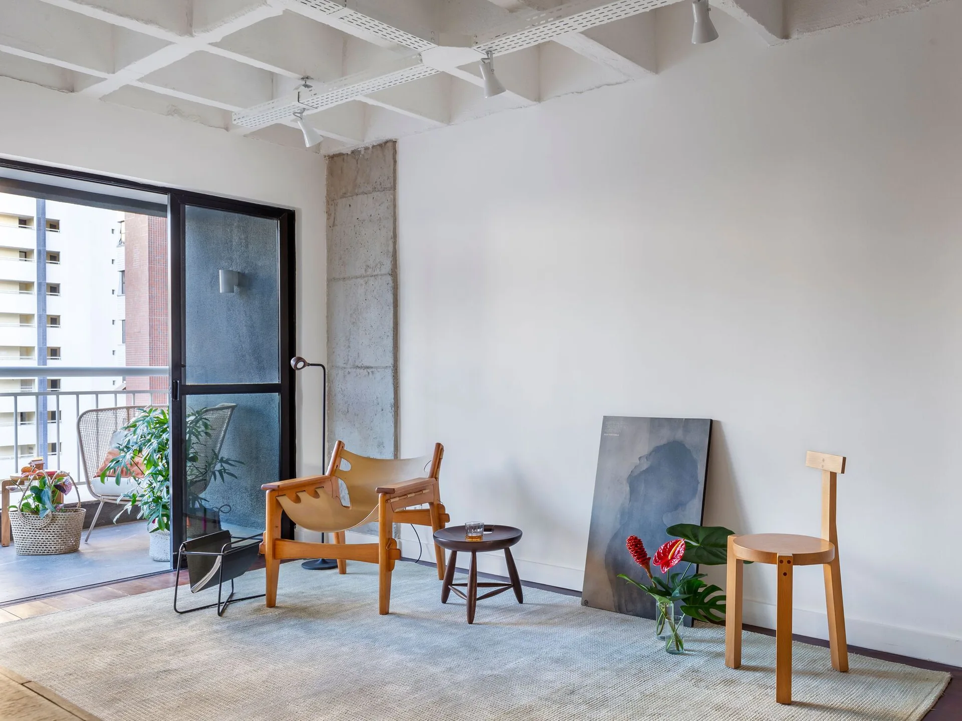A minimalist sitting area with a leather lounge chair, wooden stool, and exposed concrete waffle ceiling next to a balcony door.