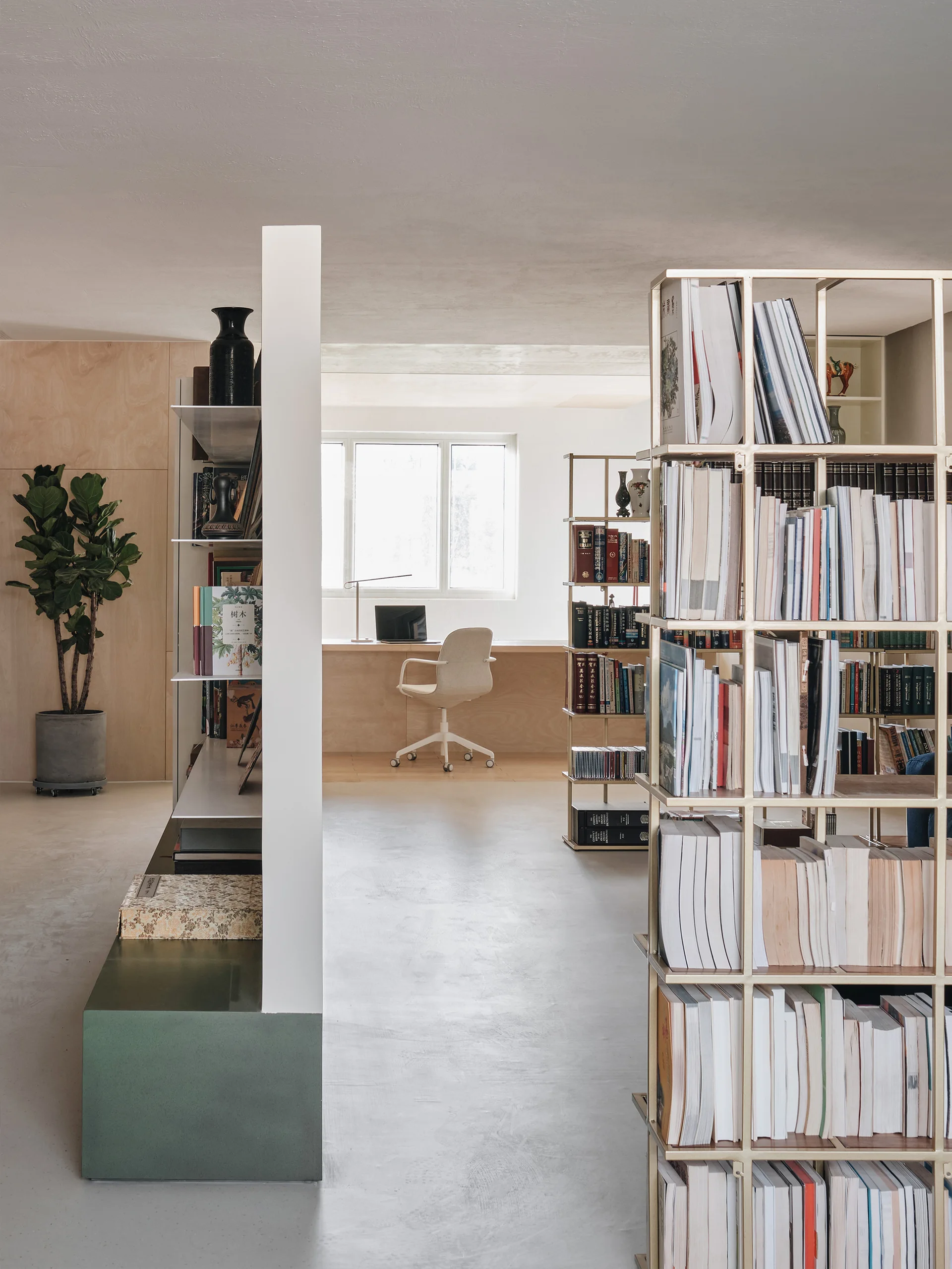 A minimalist home library features a built-in plywood desk by a window, a white rolling chair, and gold metal open bookshelves filled with books.