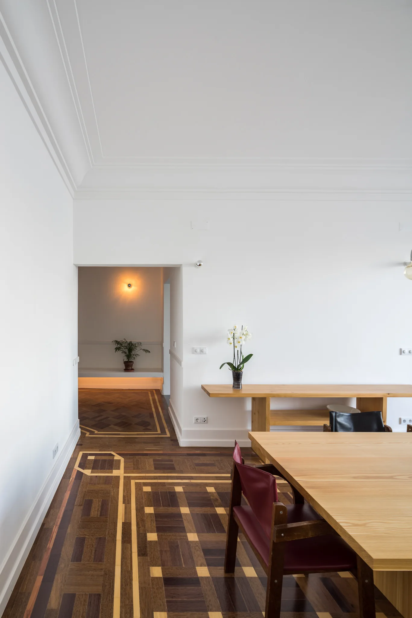 A minimalist dining room features a light wood table, red chairs, and a long wood console on detailed geometric inlaid parquet flooring.